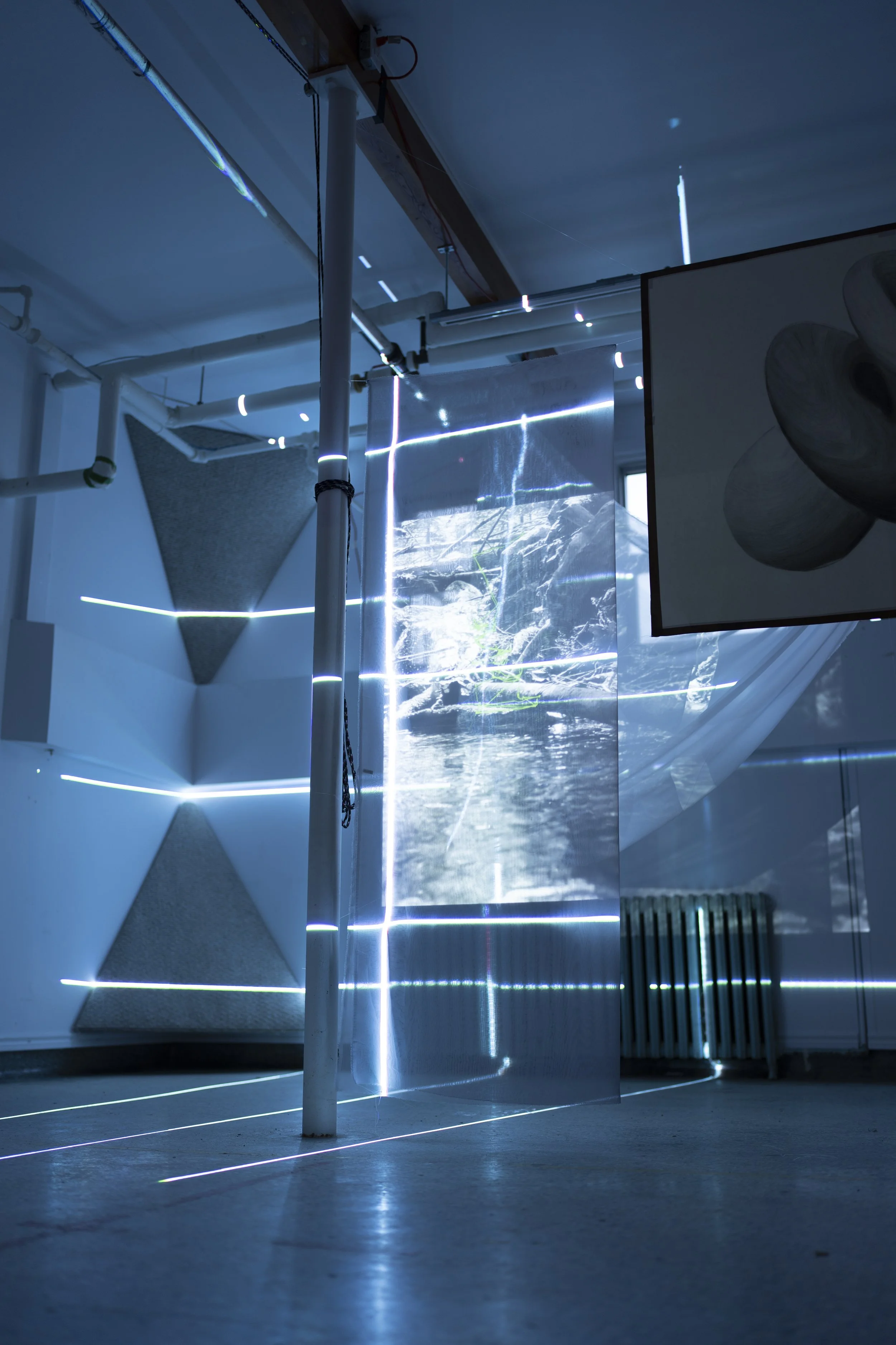 An art installation with blue LED lights on white walls, a projection of a river scene on a semi-transparent screen, and geometric textured panels, creating a futuristic atmosphere.