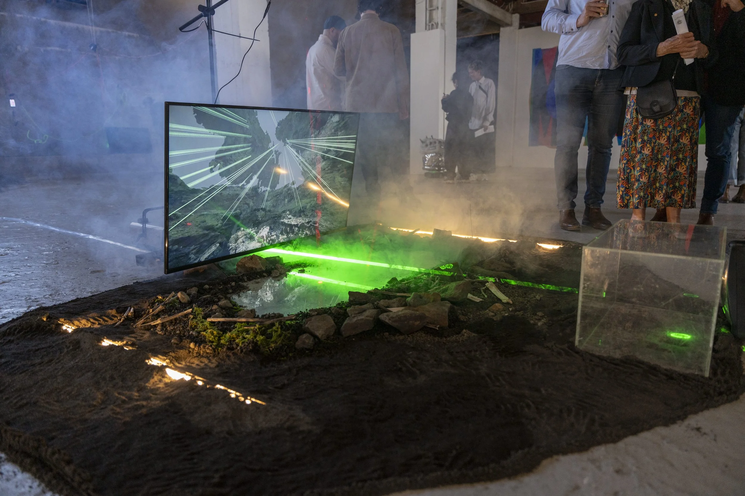 A diorama with a simulated earthquake scene, including a large screen displaying a lightning storm, a model of a crack in the ground with rocks, dirt, and green laser lights, and a group of people observing in the background.