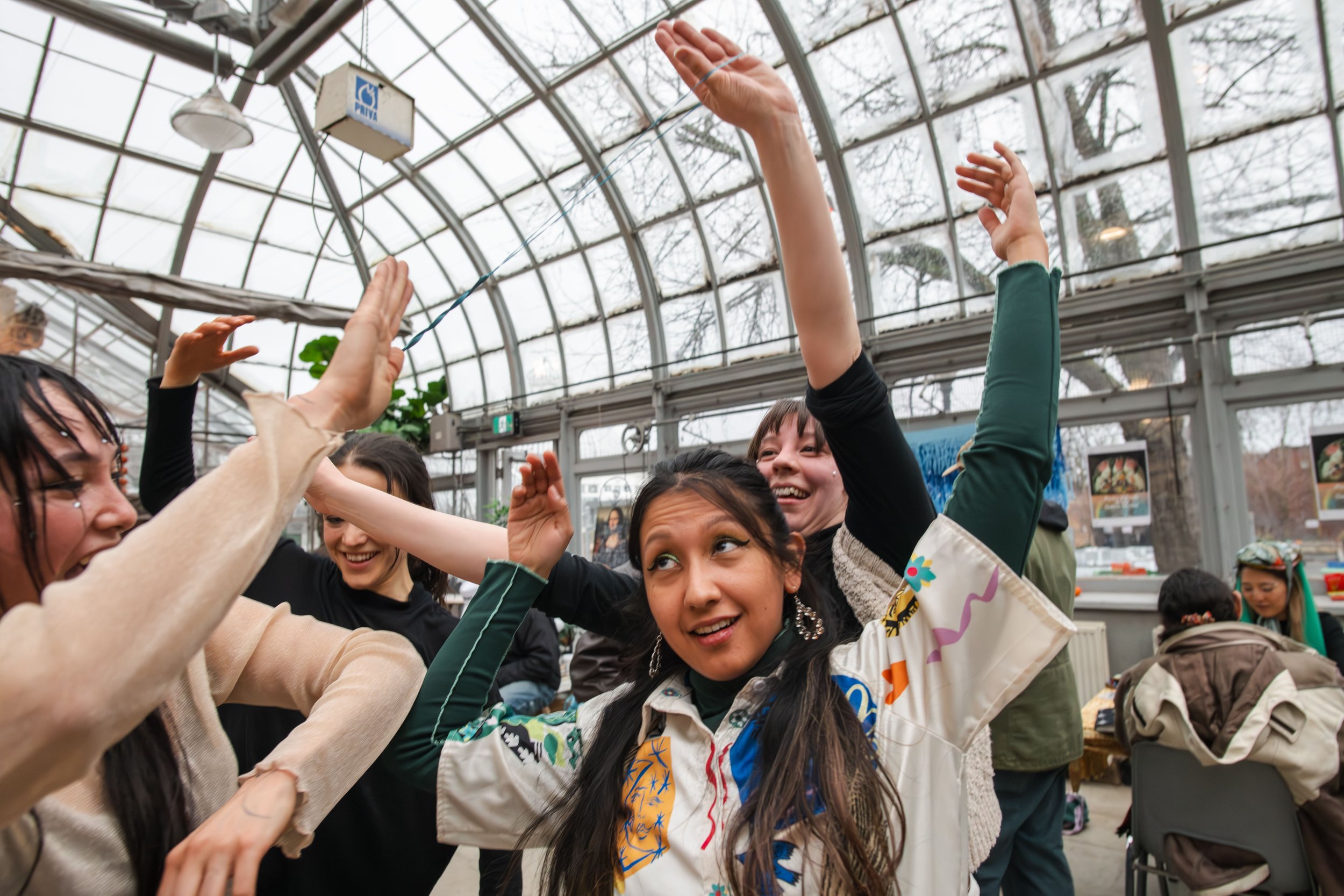 Group of young women laughing and celebrating inside a greenhouse with large glass windows and trees outside.