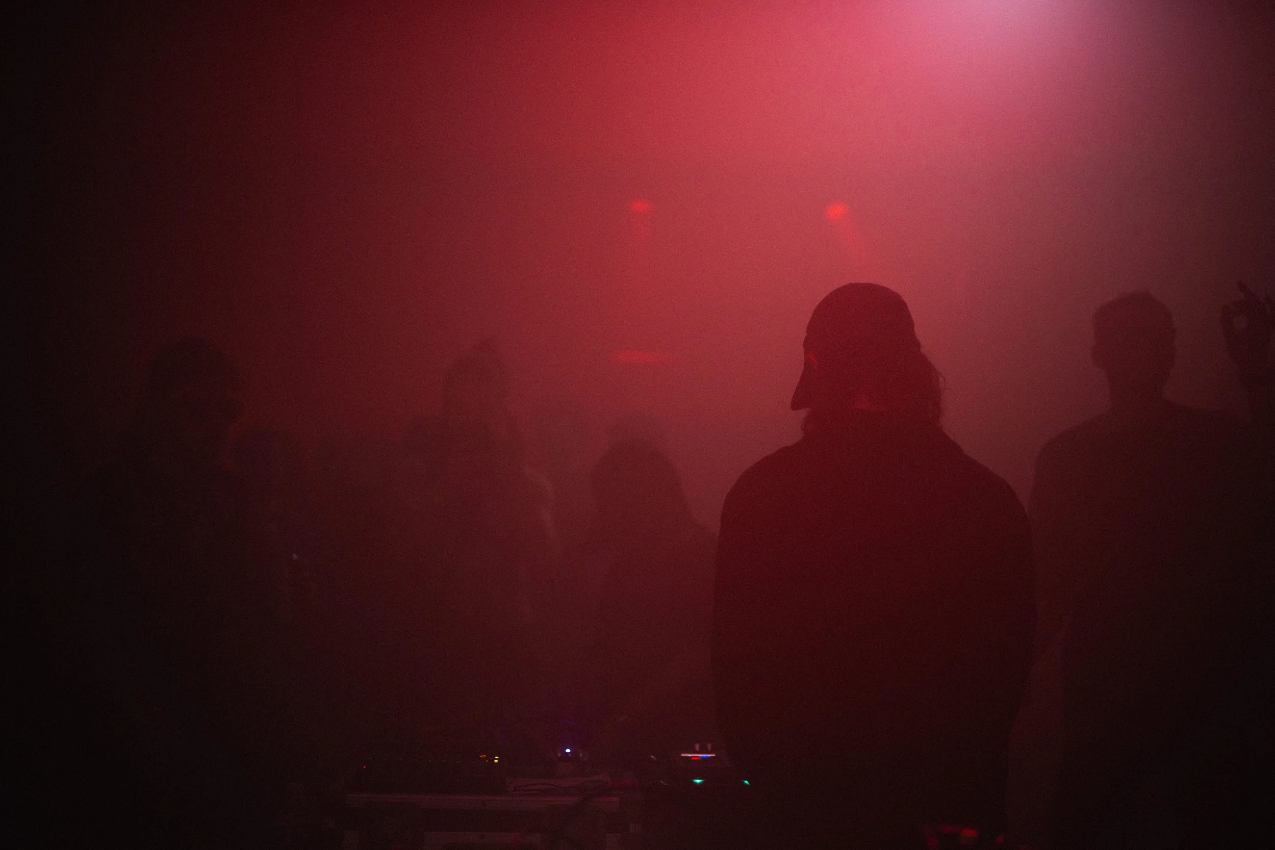 People dancing in a dark, foggy venue with red lighting, some wearing glasses, and a person with long hair in the foreground.
