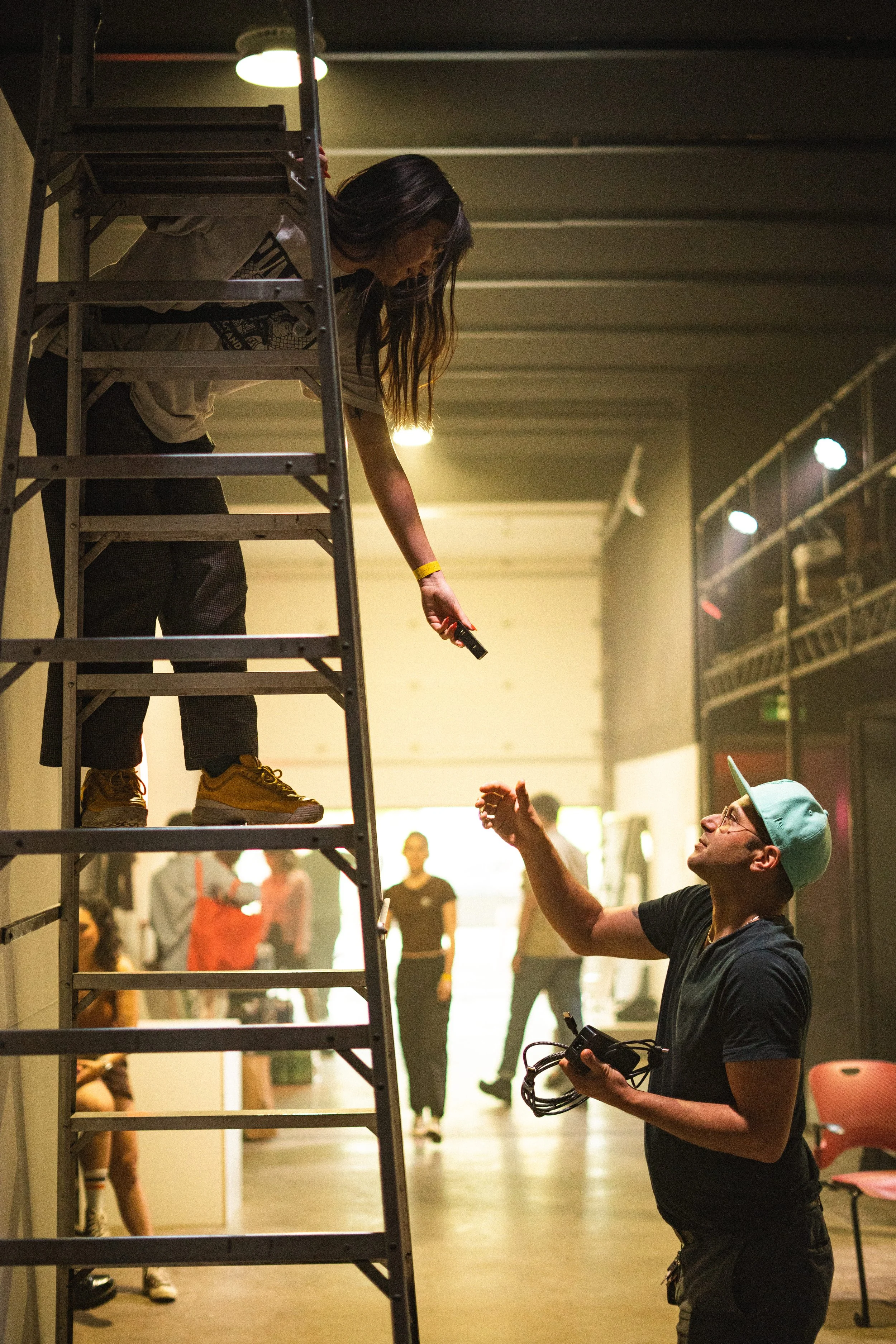 Two people, one on a ladder and one standing below, exchanging a handgun, in an indoor setting with others in the background.