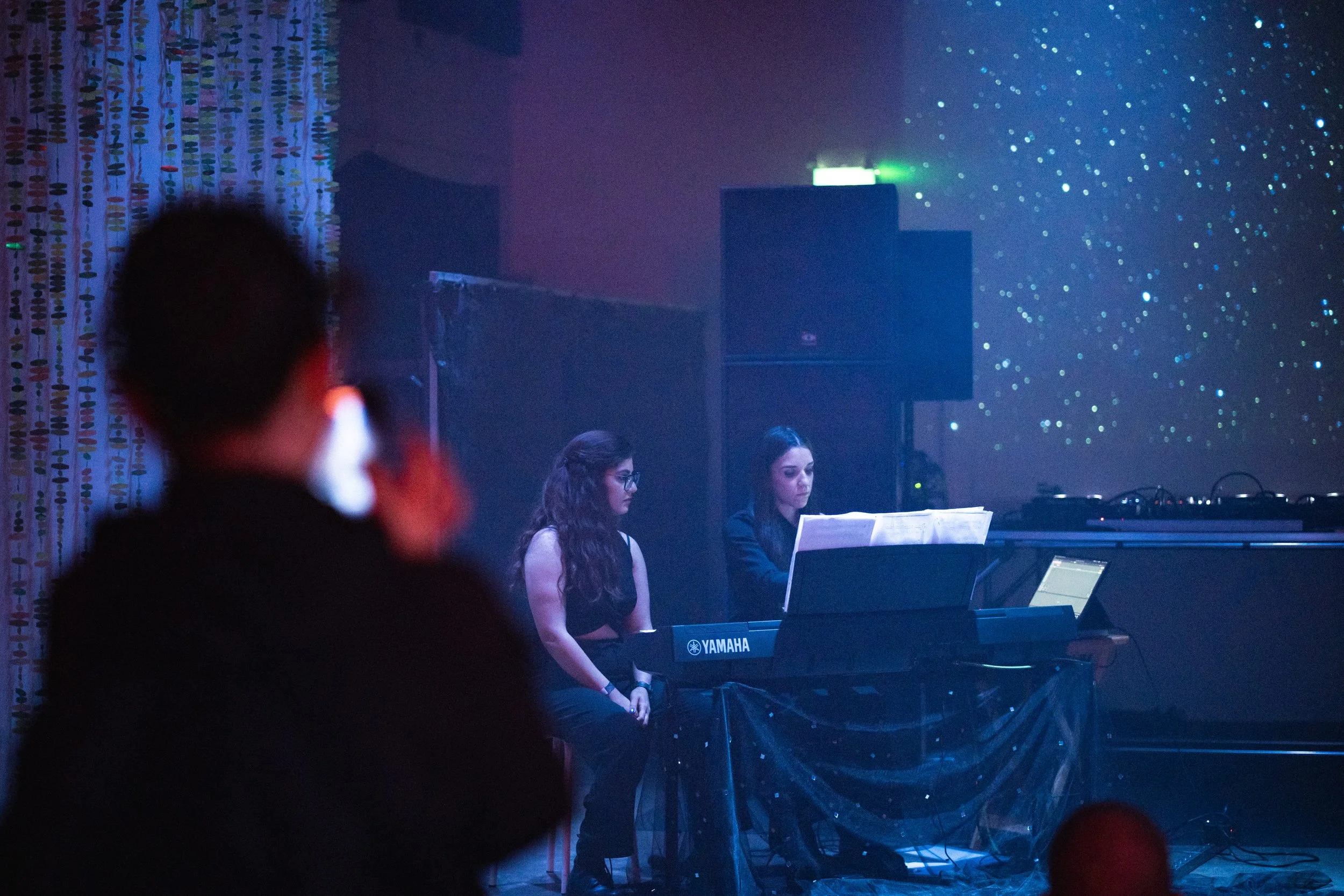 Live musical performance featuring two women sitting at a Yamaha keyboard on a dimly lit stage, with a person blurred in the foreground and a backdrop of starry lights.