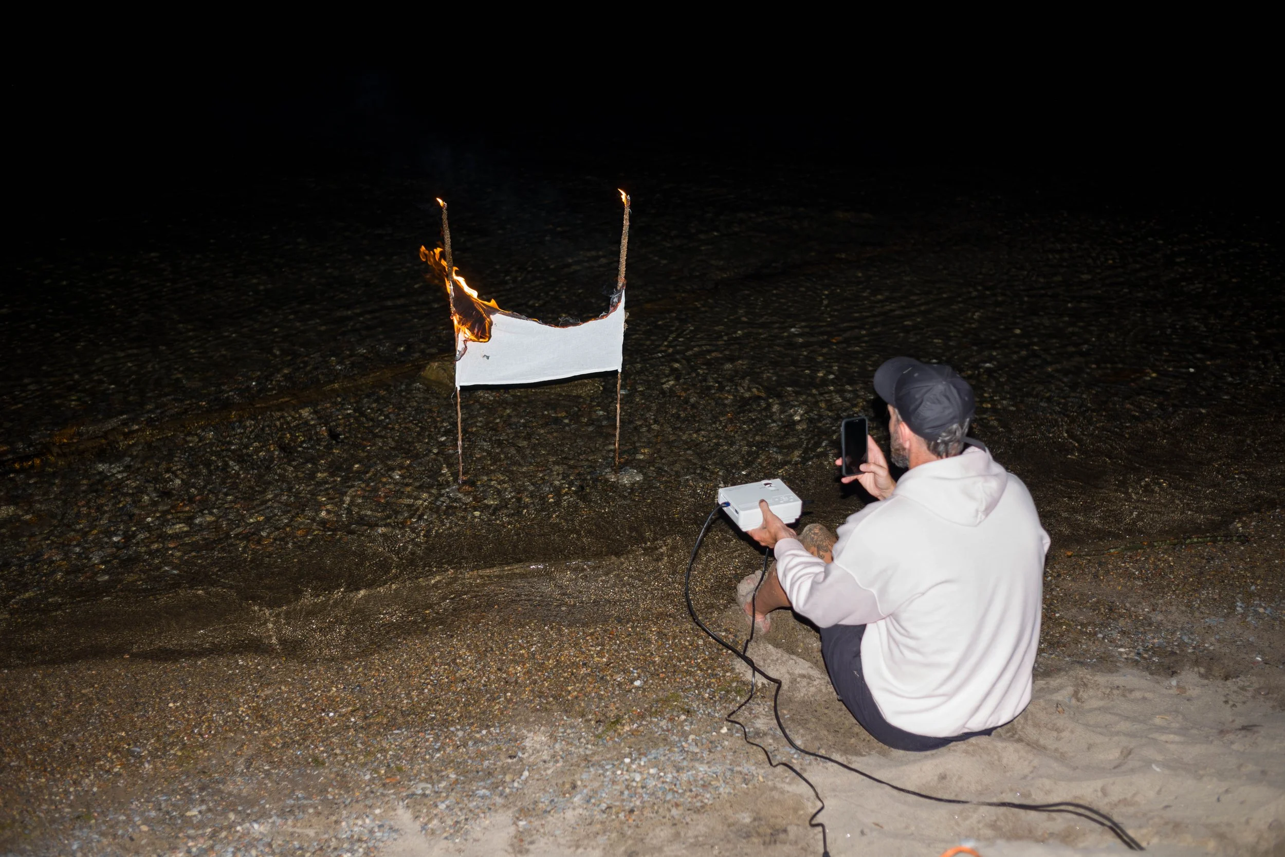 A person kneeling on a beach taking a photo of a burning white paper. The person is holding a smartphone and equipment connected with a cable, wearing a white hoodie and black cap, at night.