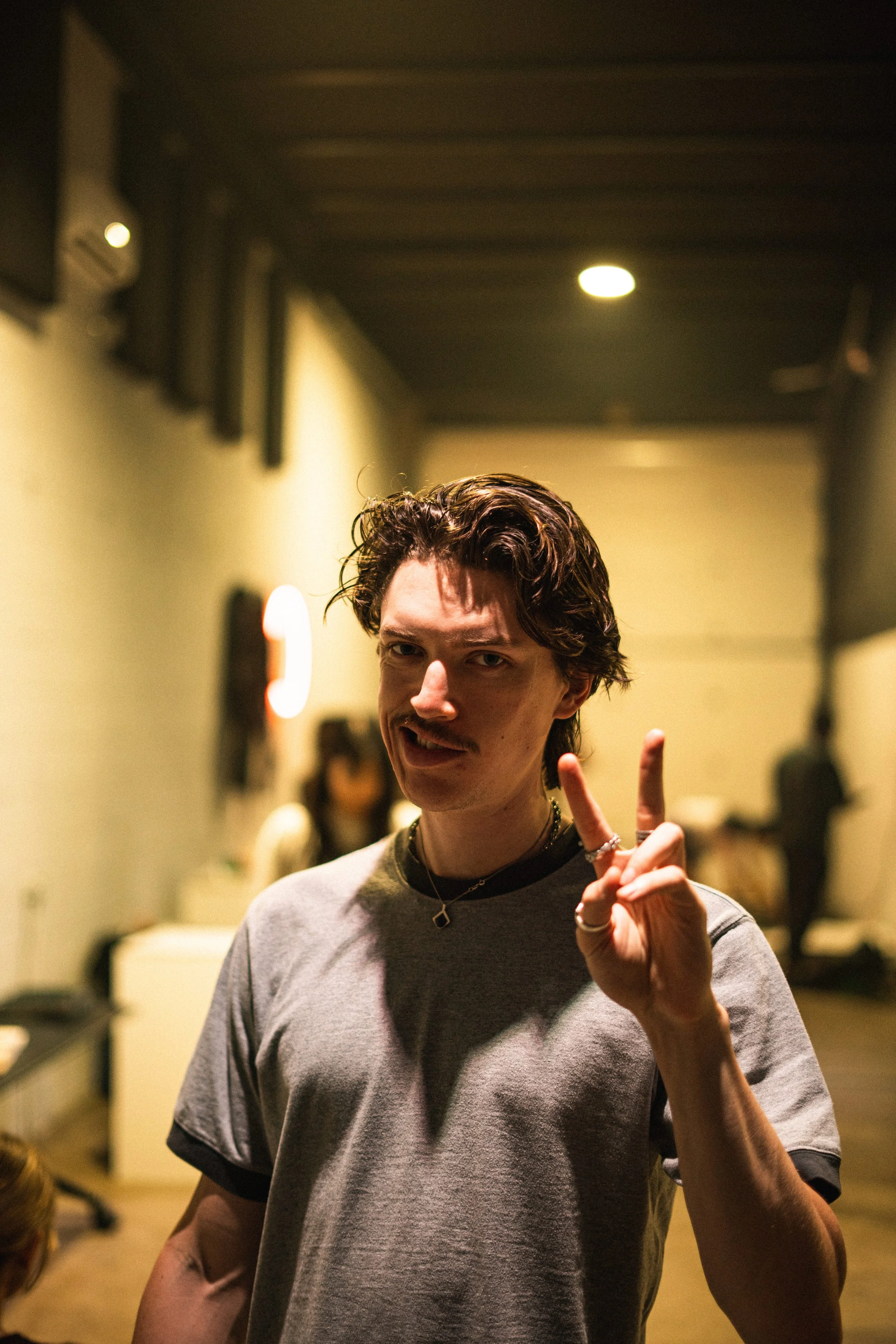 A young man with dark hair and a mustache making a peace sign with his left hand in an indoor setting with warm lighting.