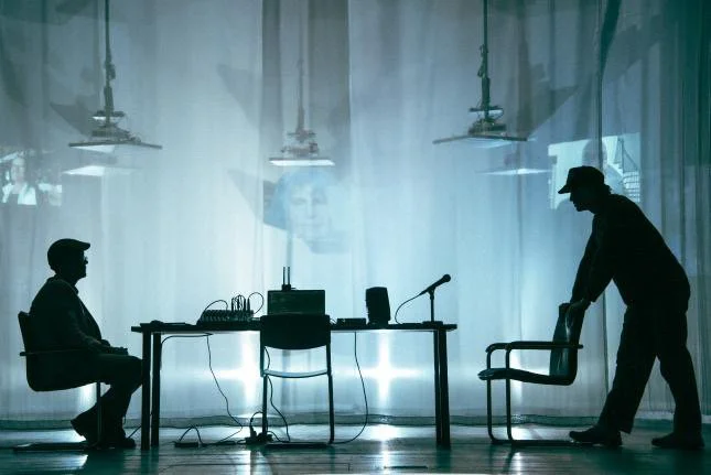 Two people are in a room with a large window, with the person on the left sitting on a chair and the person on the right moving a chair. There is a table with audio equipment and microphones, and curtains are drawn across the window.