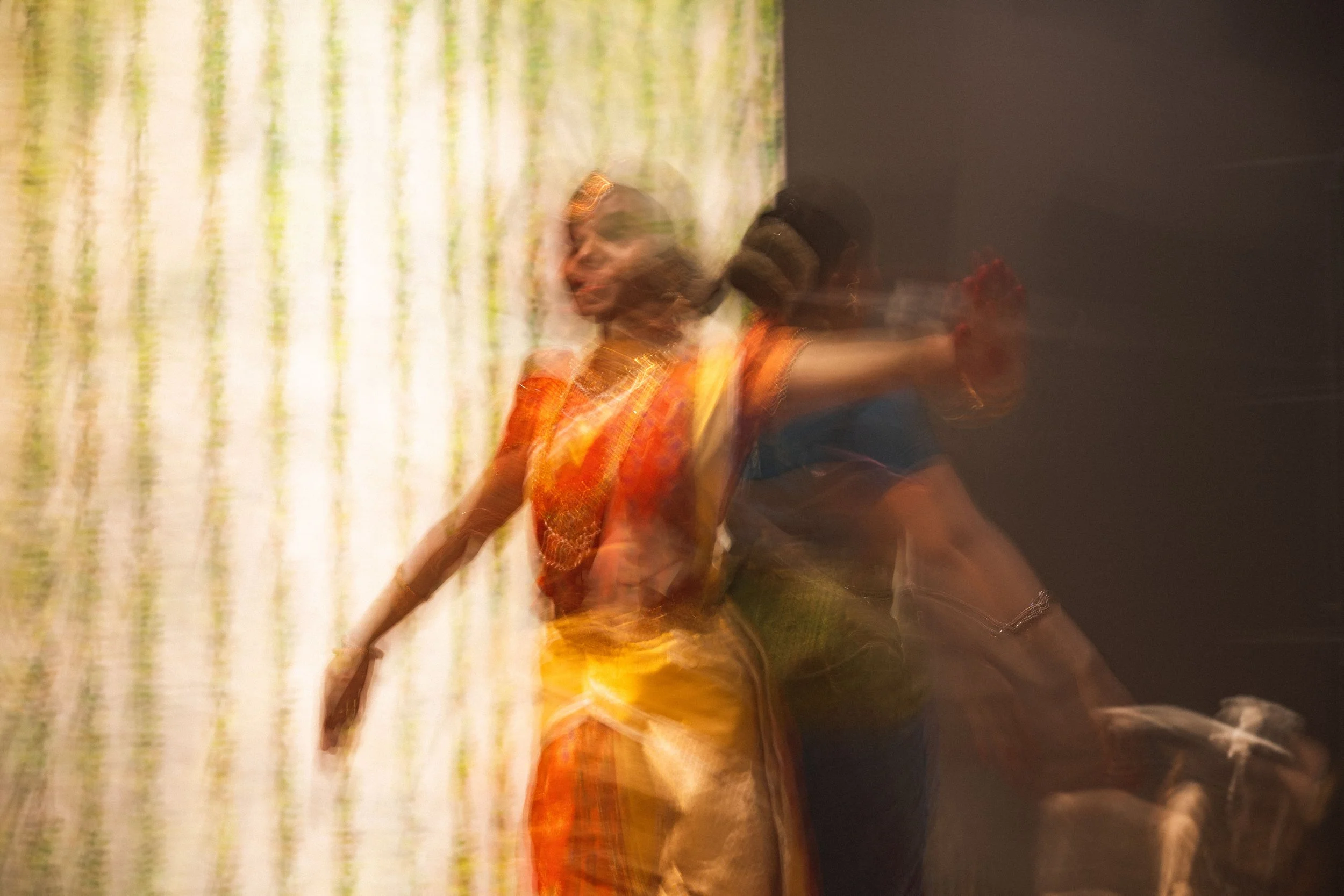 Blurred image of a woman wearing traditional colorful attire, dancing or moving against a patterned background.