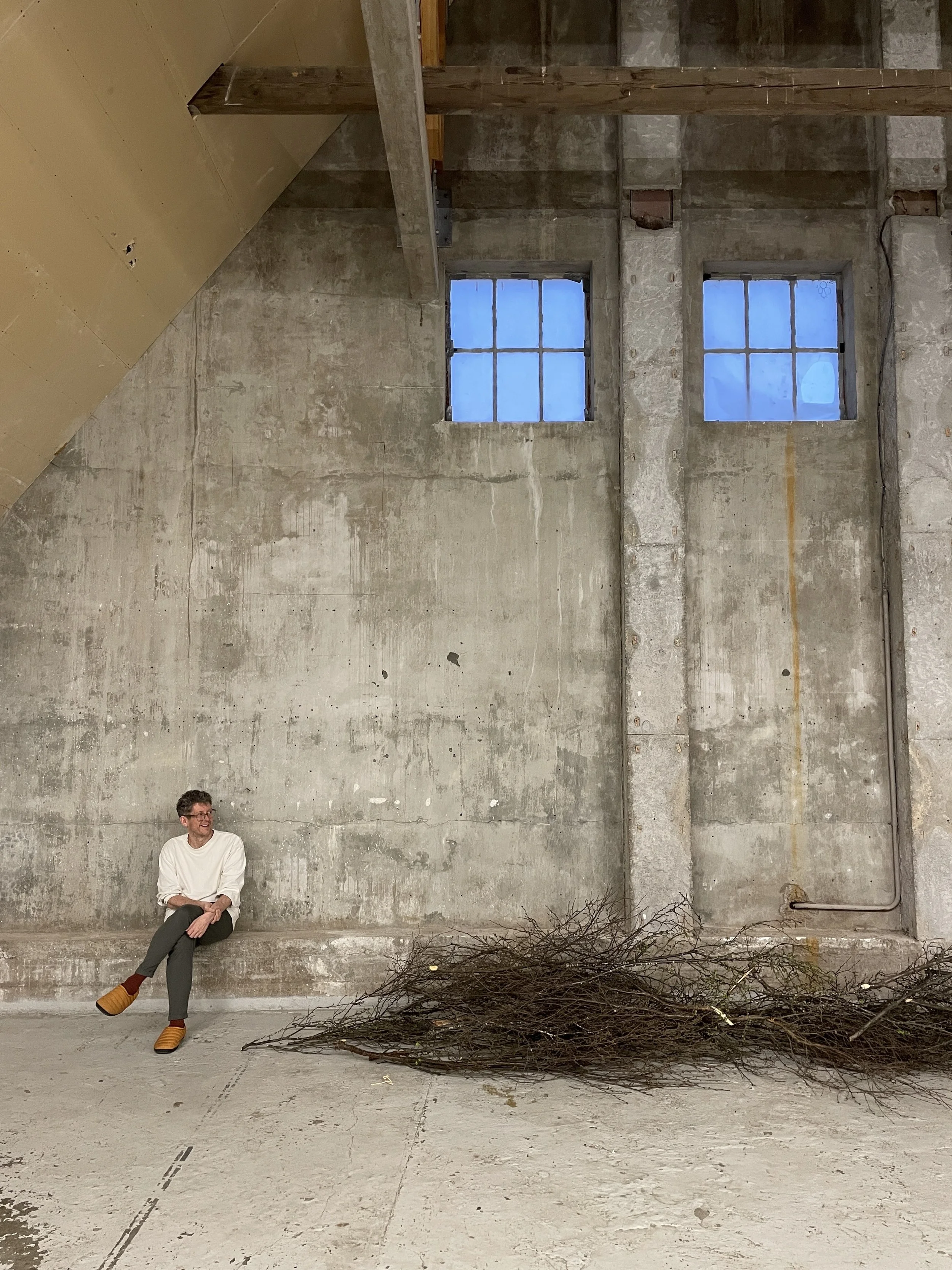 A person sitting on a concrete ledge inside an unfinished building with two small windows, a pile of bare branches on the floor, and exposed wooden beams.