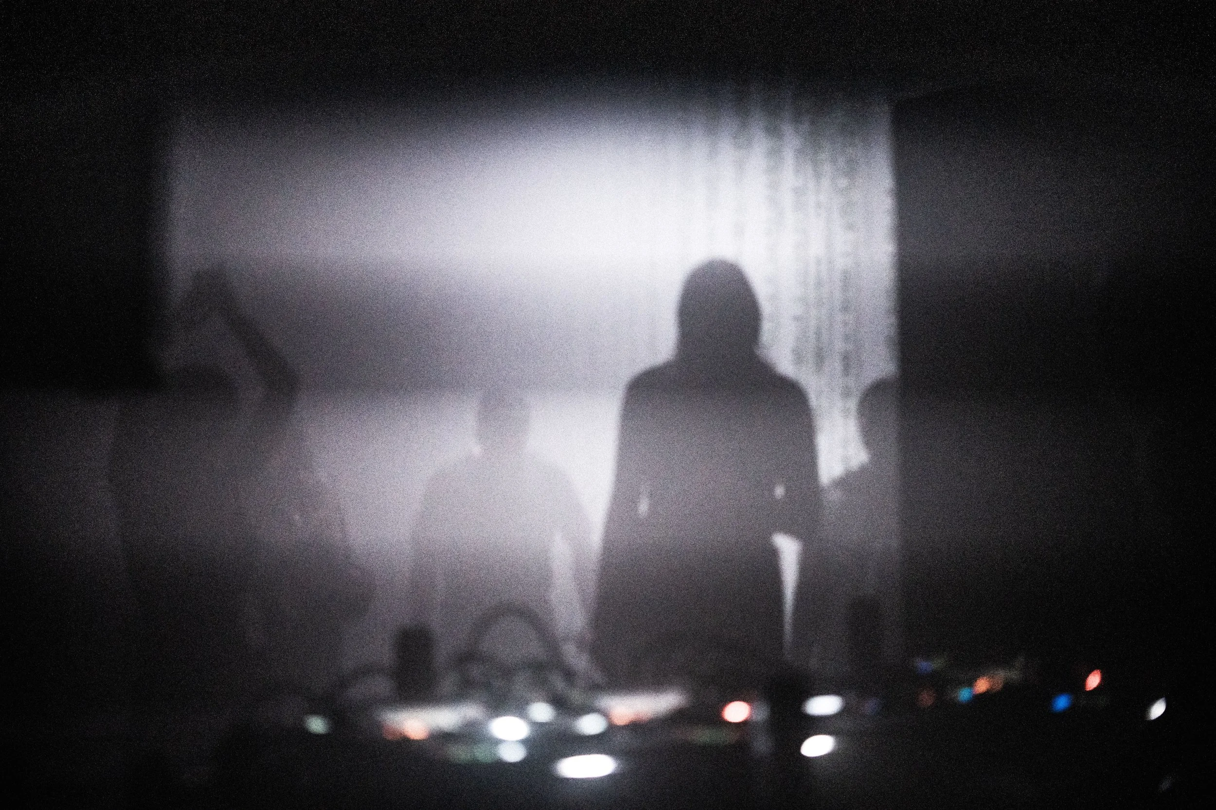 Silhouettes of people, including a DJ, in a dark setting with a bright white beam of light and abstract background patterns.