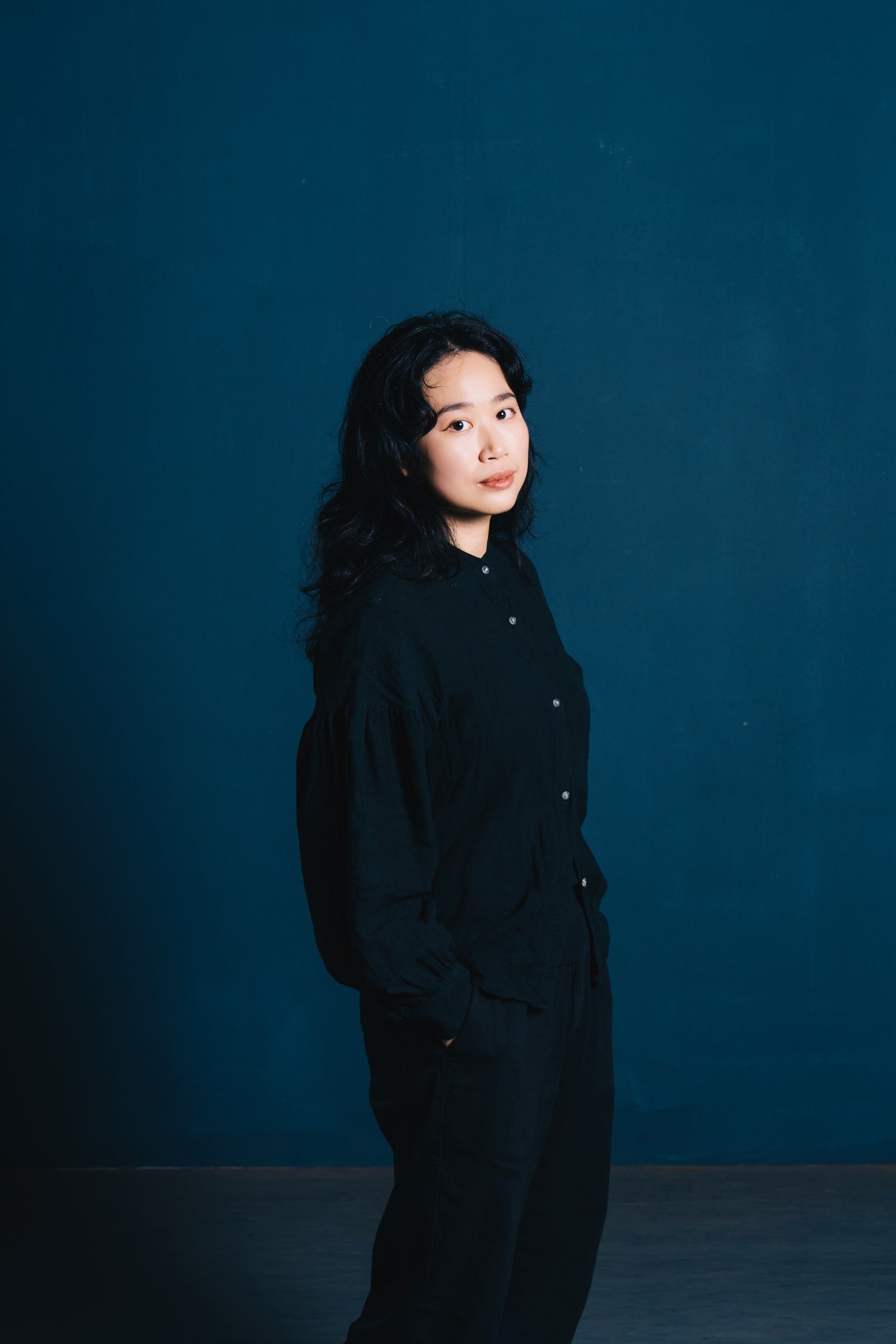 A woman with long dark wavy hair standing against a blue background, wearing a black button-up shirt and black pants.