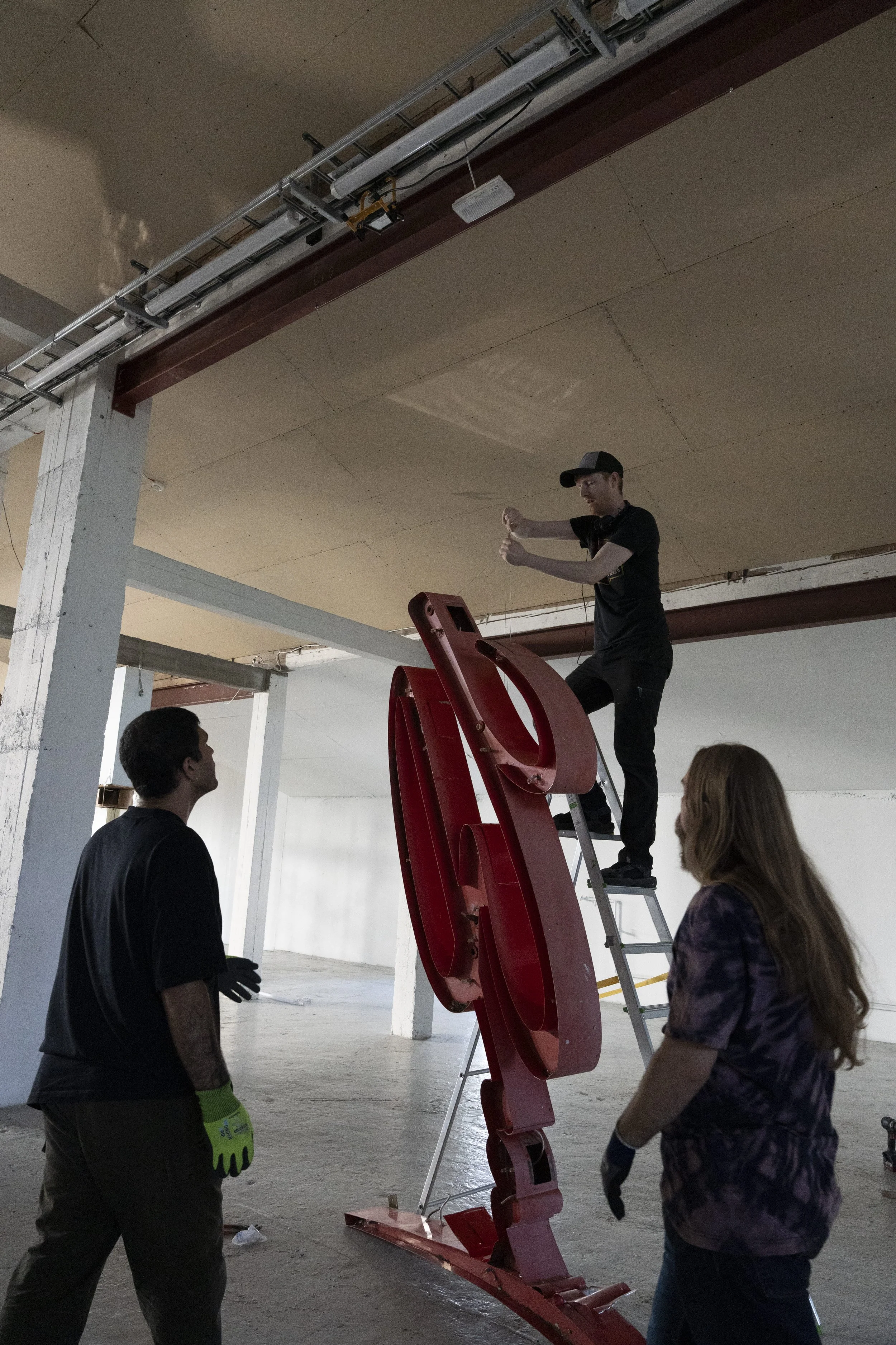 Three people working together in an industrial warehouse, assembling or installing a large red neon sign, one person standing on a ladder, two others on the ground.