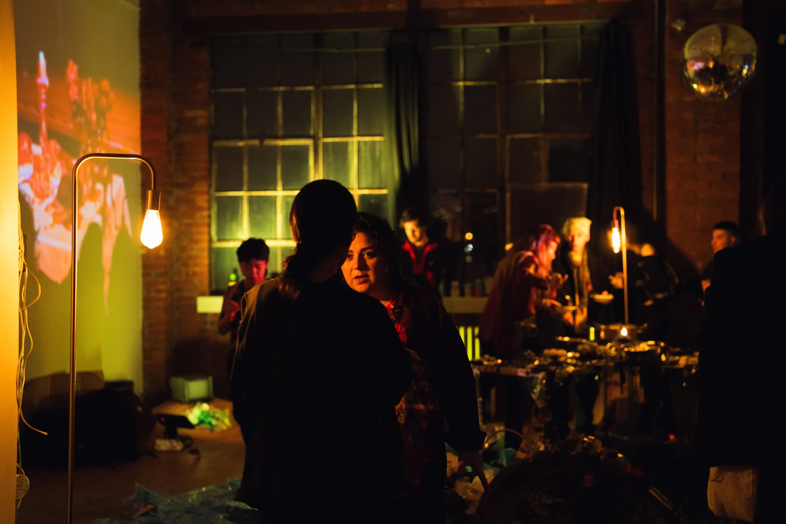 People at a dimly lit social gathering with warm lighting, a large window, and a table with food or presents.