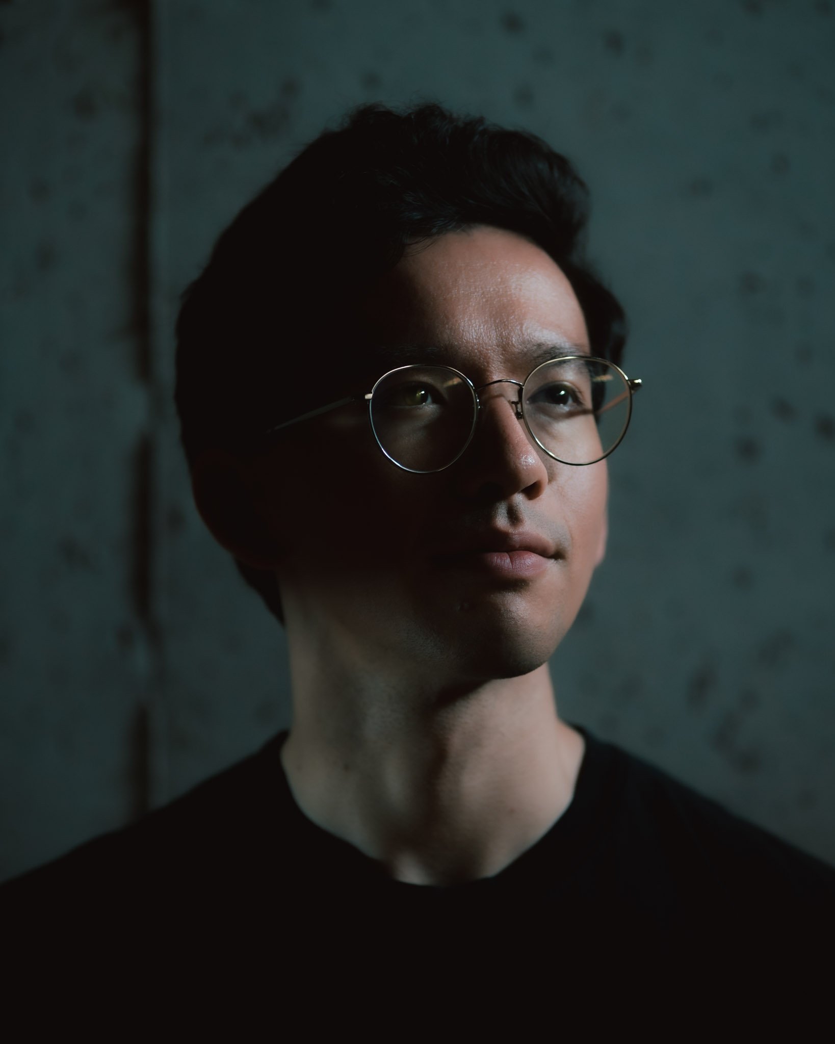 Portrait of a young man with glasses, looking slightly upward, in low lighting with a dark background.