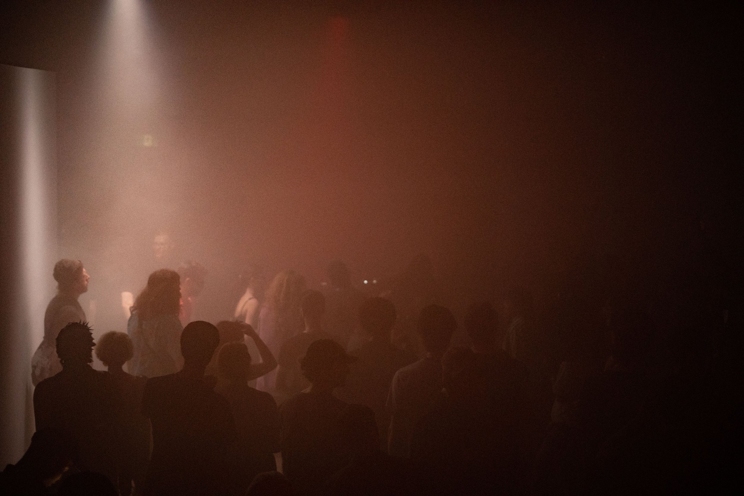 People standing in a dim indoor space with mist or smoke, illuminated by soft pinkish light from the left, facing a stage or focal point outside the frame.