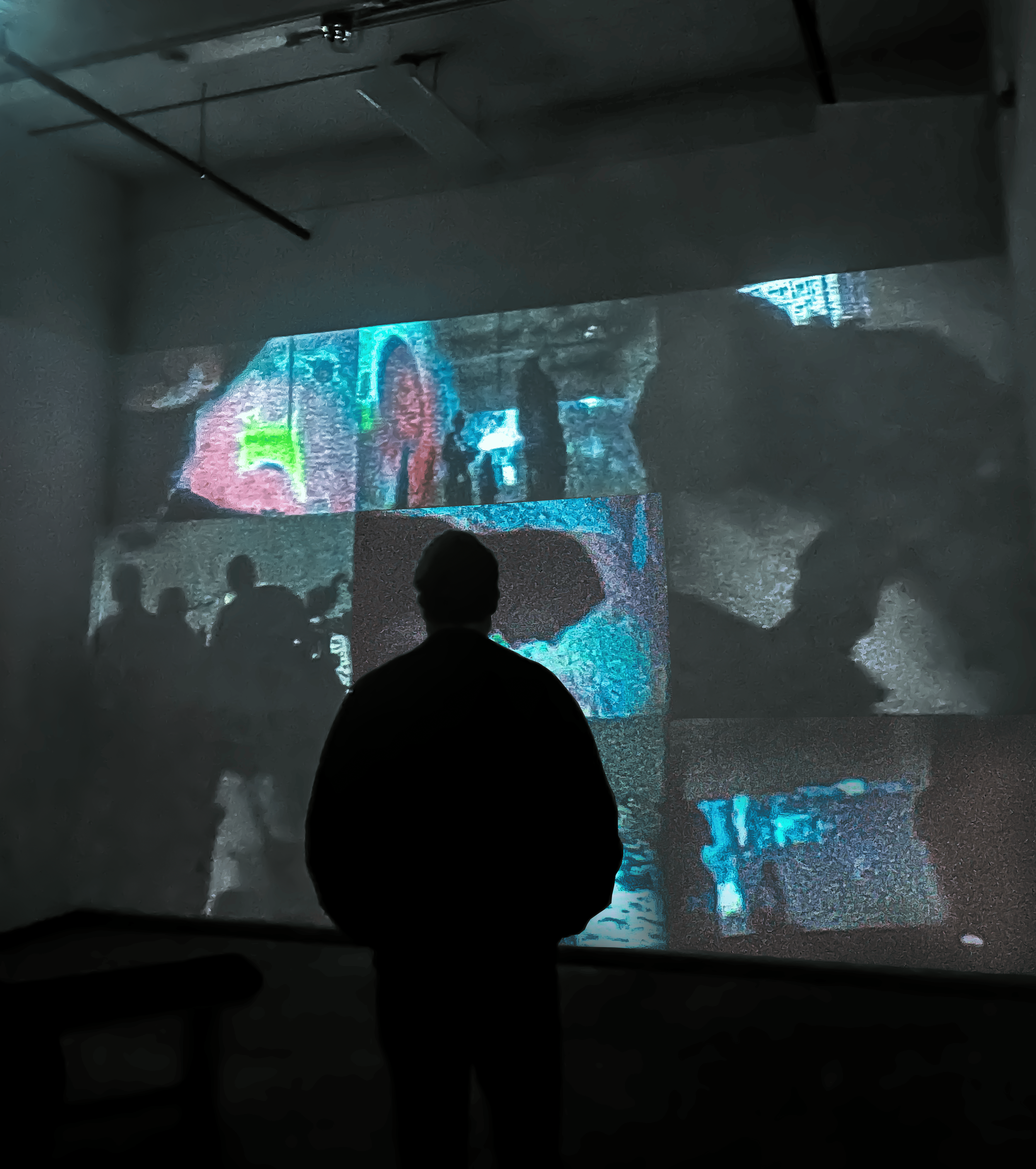 Person standing in front of a large digital or projected artwork with multiple screens displaying abstract images and silhouettes of people.