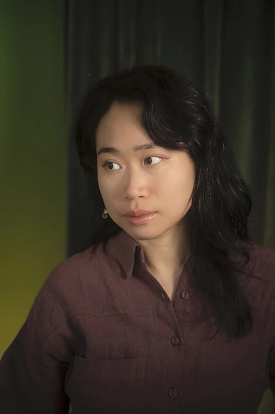 A woman with long dark hair looking to her left, wearing a maroon button-up shirt, standing in front of a green and dark background.