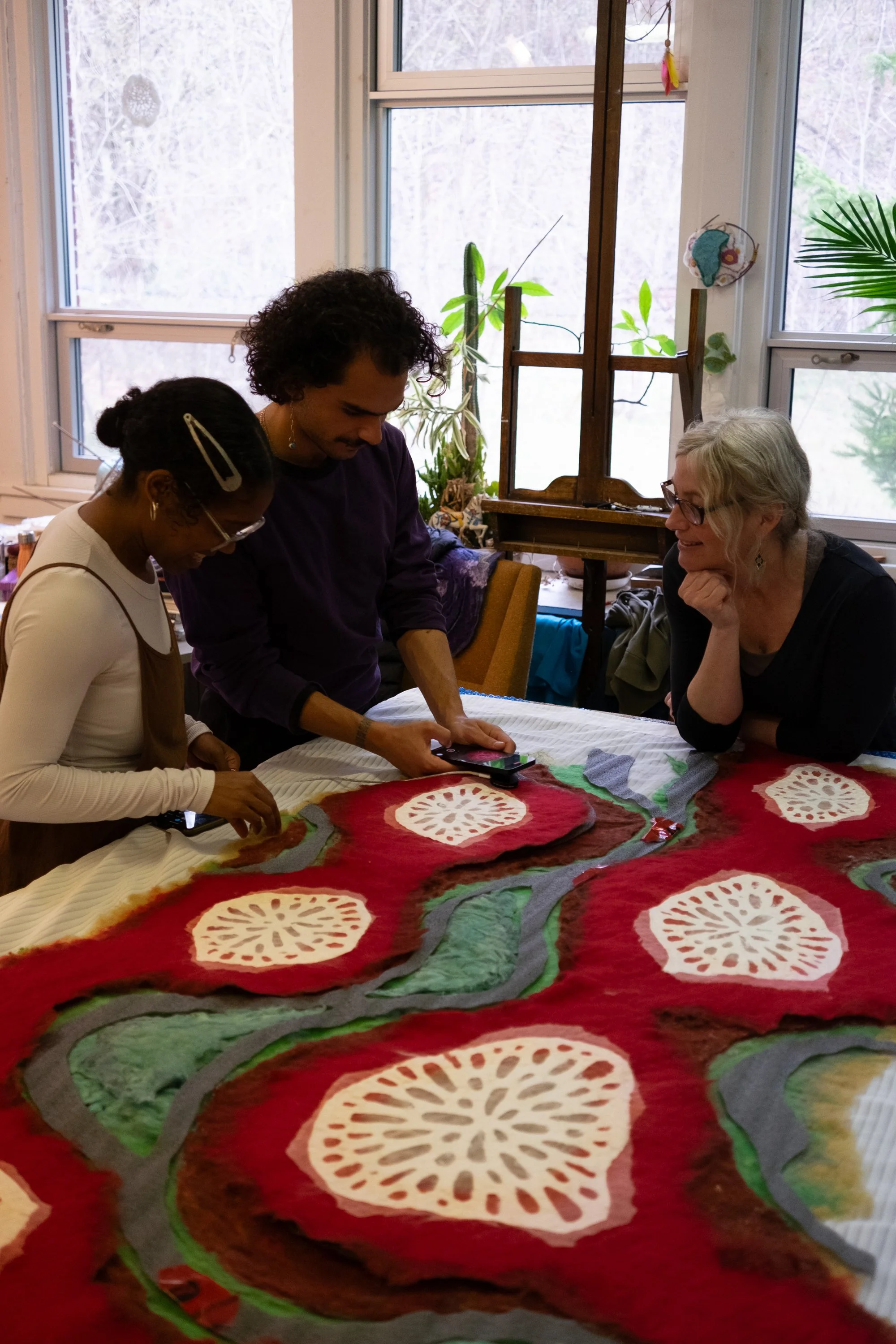 Three people gathered around a table, examining a colorful textile with organic shapes and patterns, in a room with large windows and indoor plants.