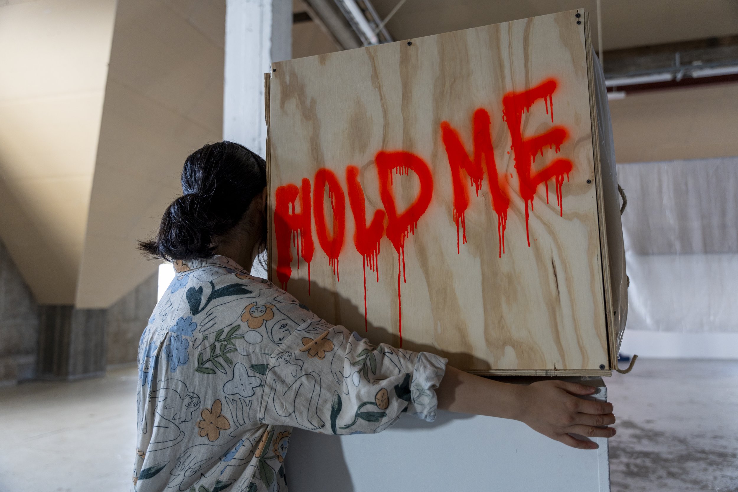A person with dark hair wearing a patterned shirt is holding up a wooden panel with the word "HOLD ME" spray-painted in red, dripping paint.