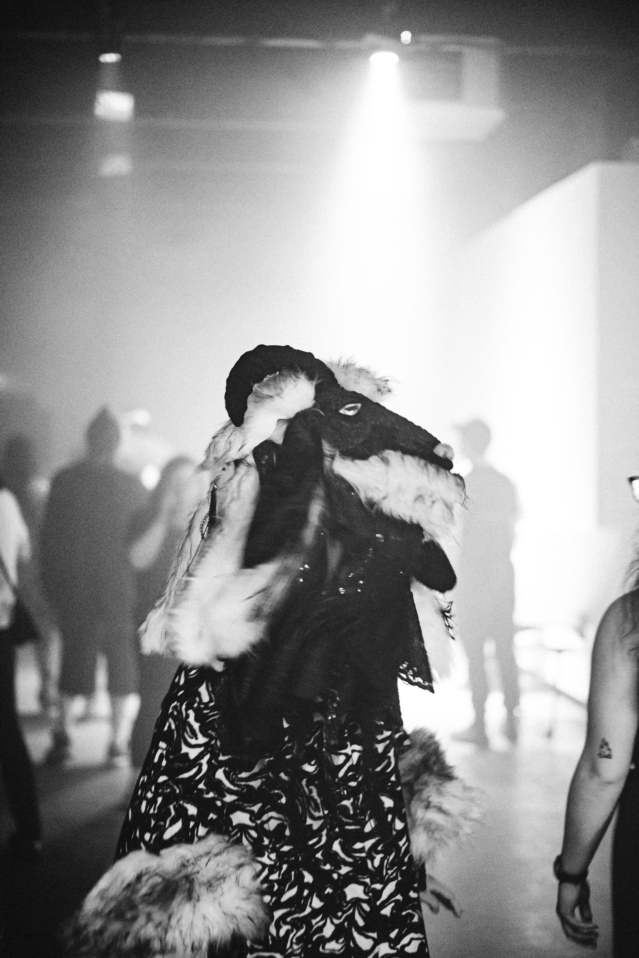 A person in a wolf costume at a dimly lit indoor event or party, with other people blurred in the background.