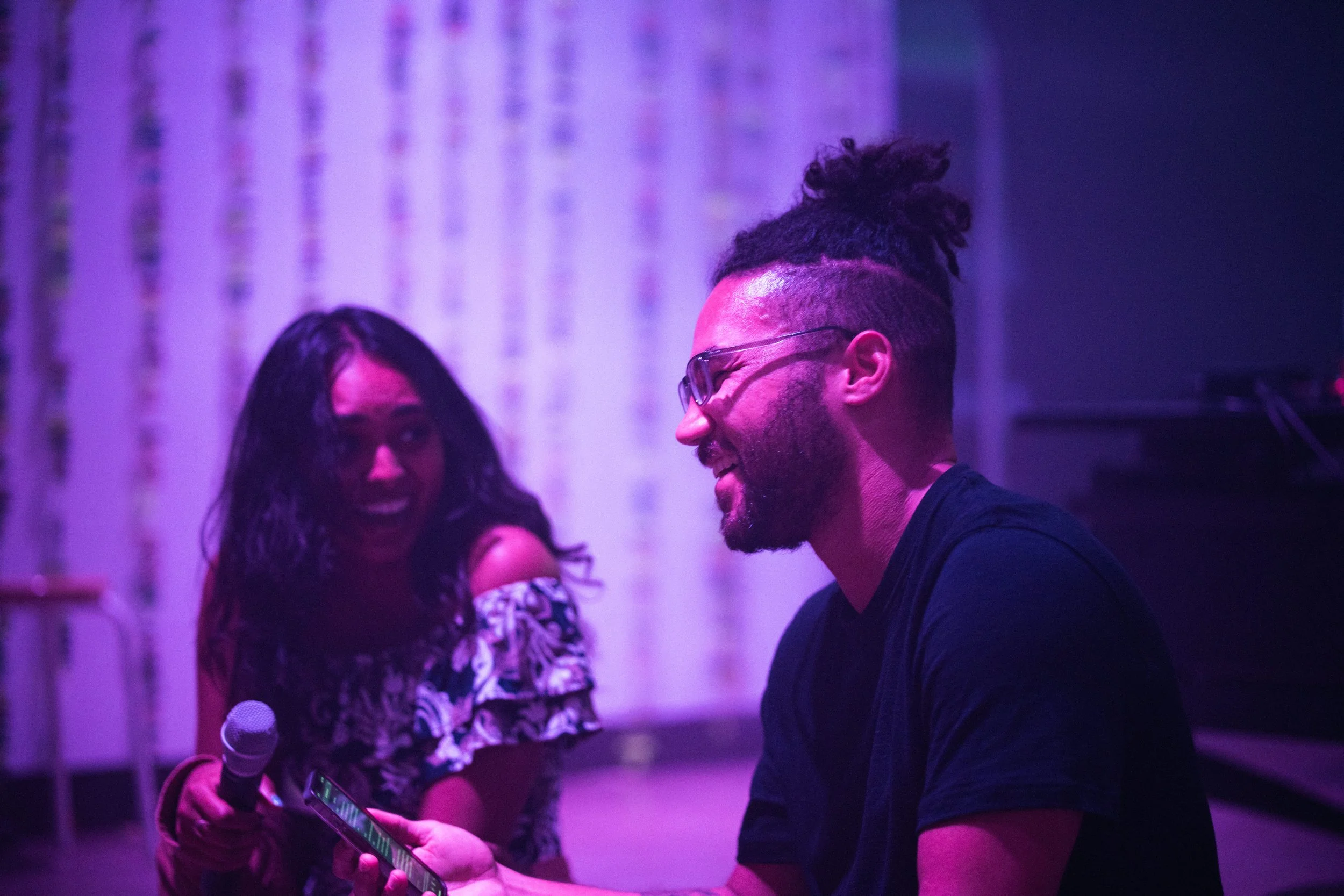 A woman holding a microphone laughs while looking at a man with glasses, who is smiling and looking at his phone, in a dimly lit room with purple lighting.