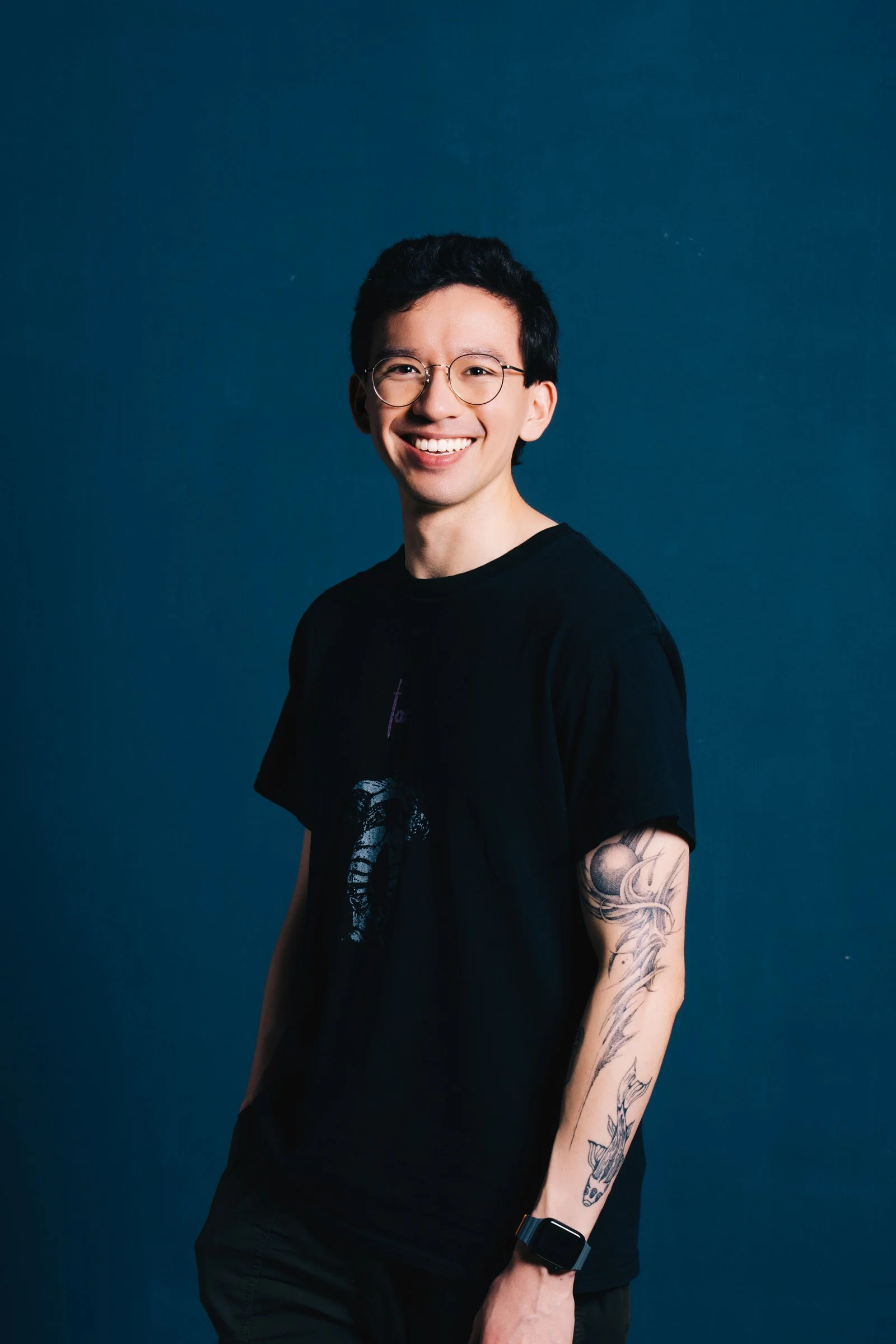 A young man with glasses, smiling, standing against a dark blue background. He has a tattooed arm and is wearing a black t-shirt and a smartwatch.