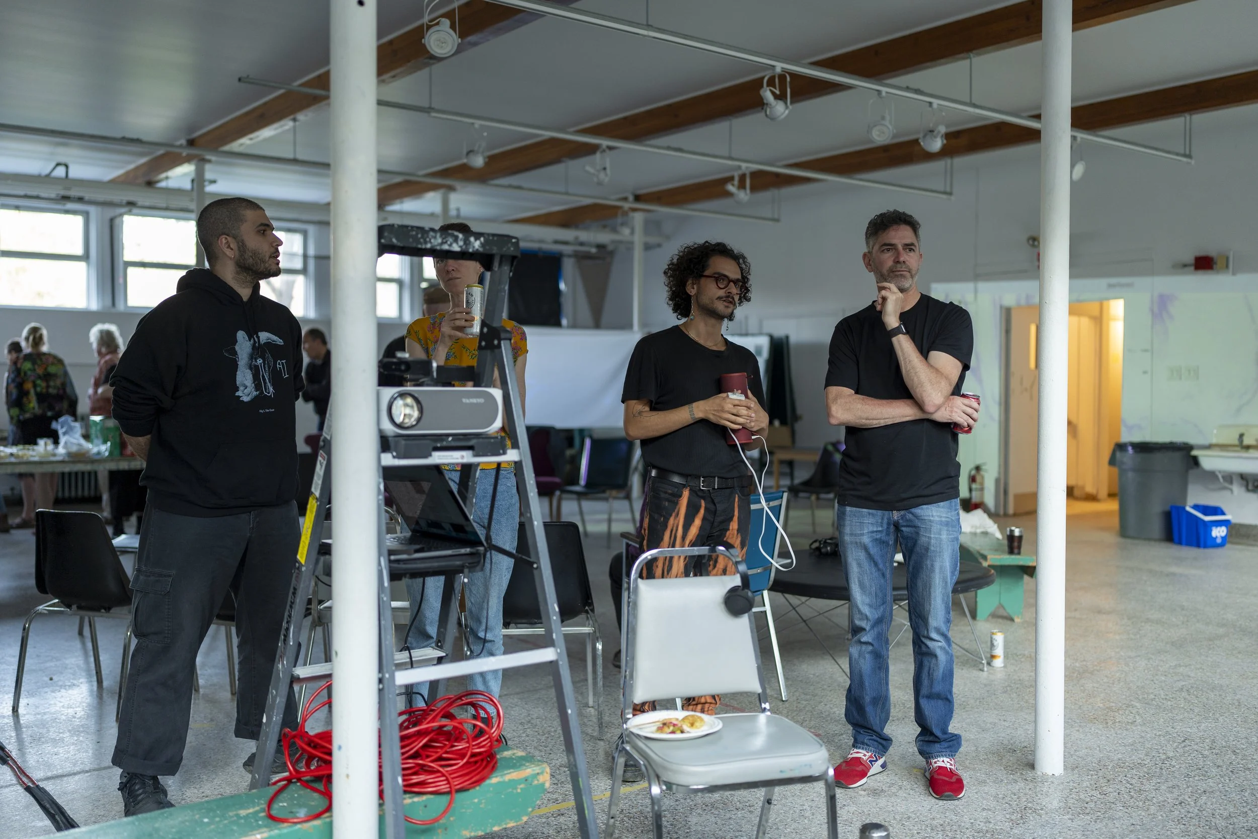 A group of five people standing and watching in a spacious room with high ceilings, white walls, and large windows. There are chairs and tables in the background, and various equipment and supplies are scattered around.
