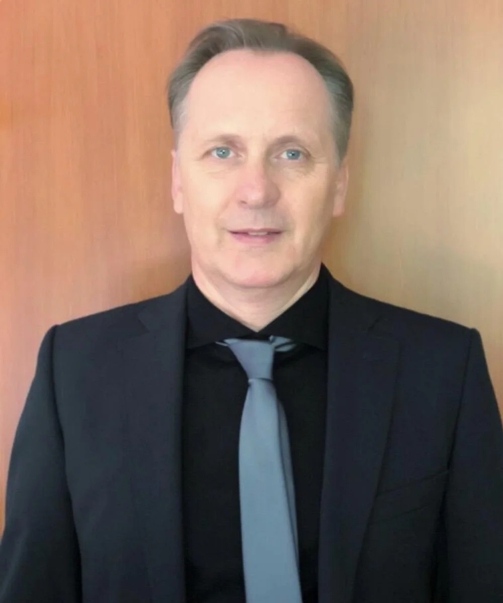 Ein Mann mit kurzen braunen Haaren in einem Anzug steht vor einer Holzwand und blickt direkt in die Kamera.