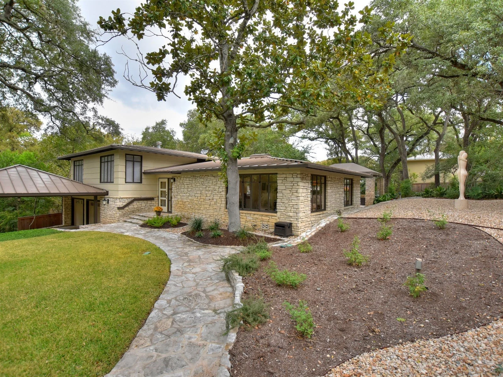 Modern house exterior with stone accents, surrounded by trees and landscaped yard, featuring a stone path and a sculpture.