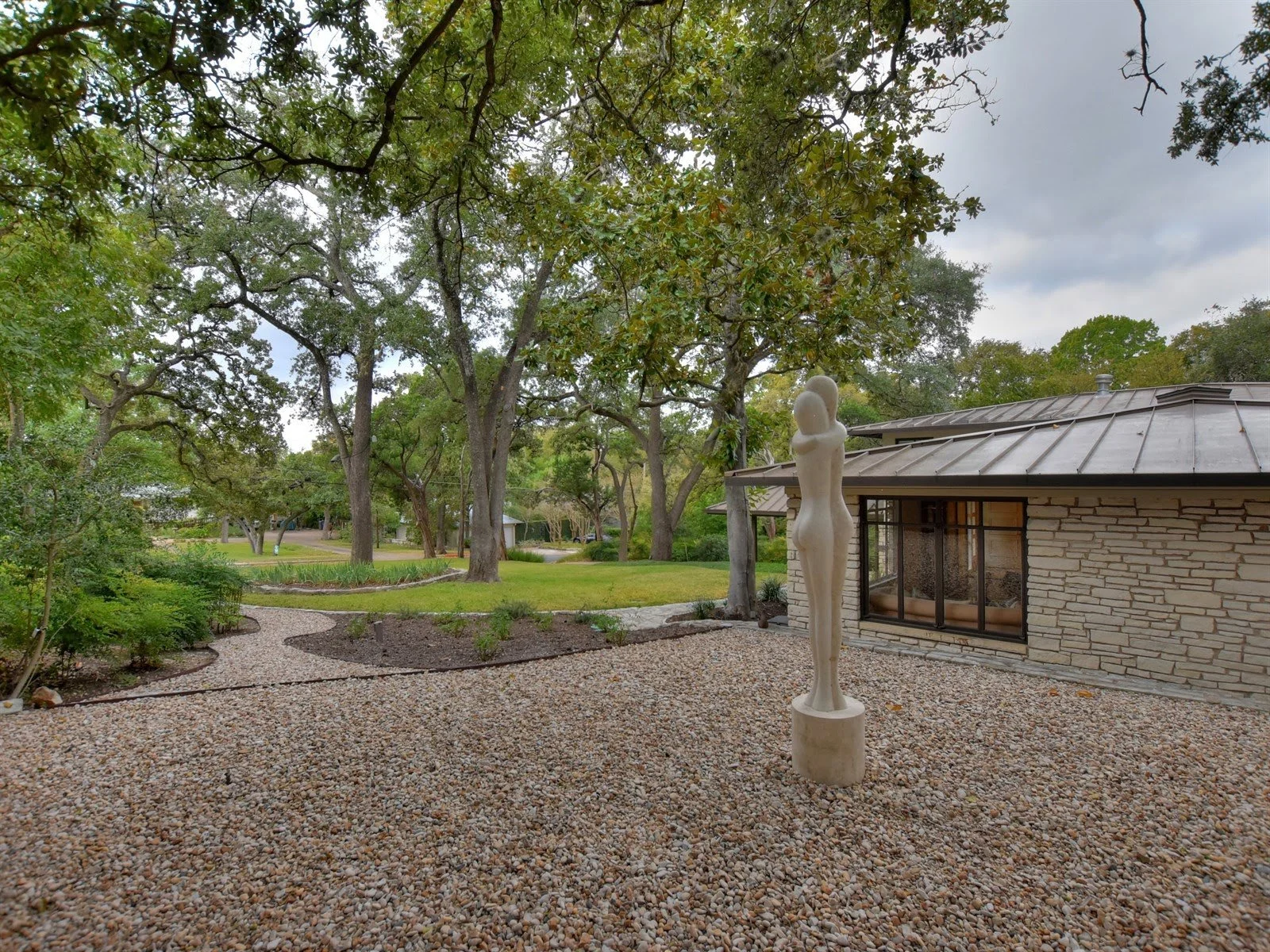 Backyard garden with a stone pathway, lush greenery, large trees, a modern stone house, and a sculpture on a gravel area.