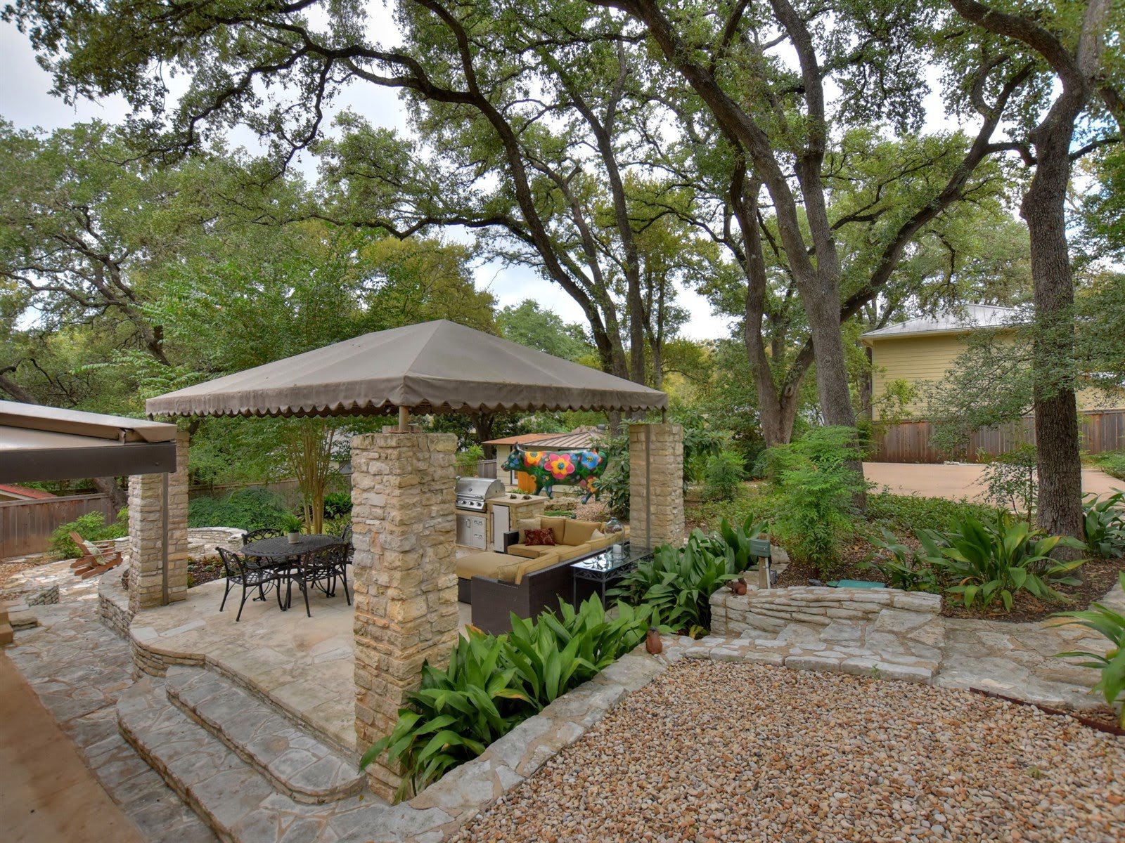 Outdoor stone patio with a covered canopy, dining table, chairs, grill, surrounded by trees and plants.