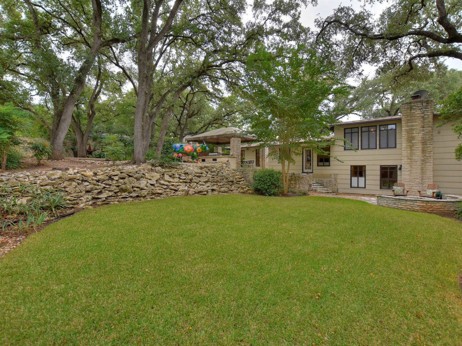 Backyard with green lawn, trees, stone wall, and a house with large windows and a chimney. Colorful sculpture visible near the patio area.
