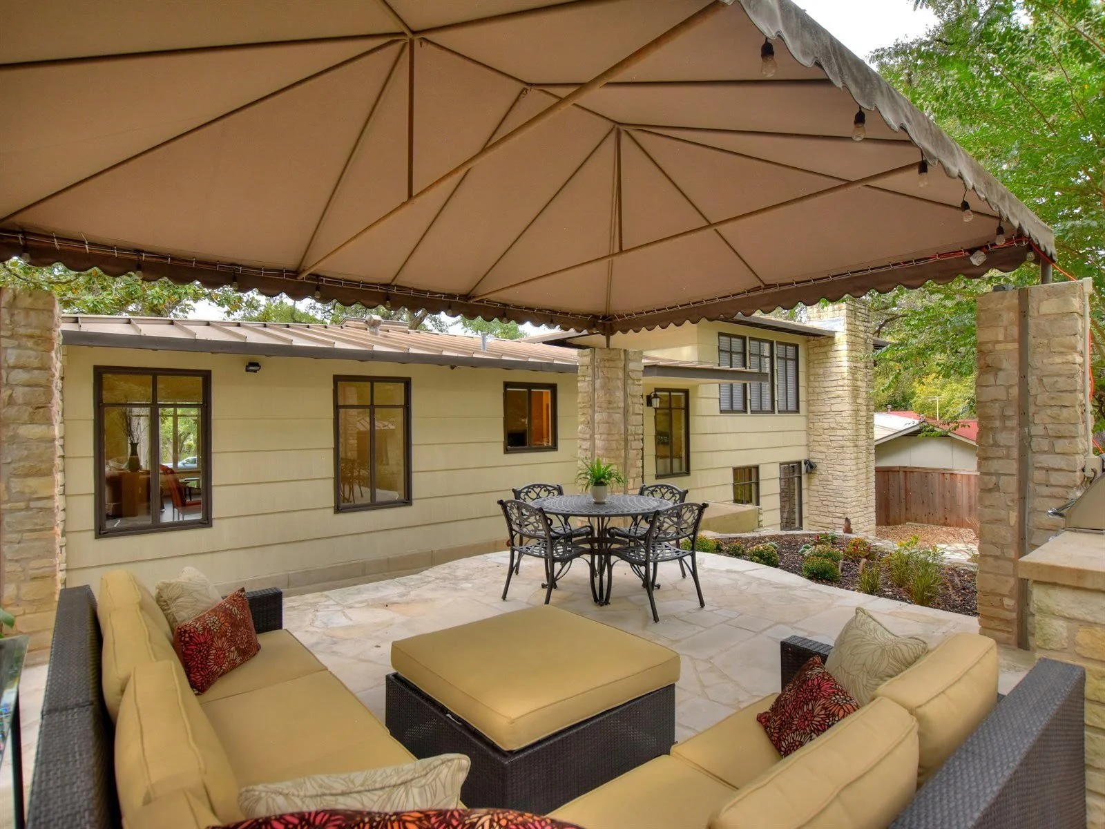 Outdoor patio area with beige cushioned seating, a metal dining table with chairs, under a canopy at a house with stone pillars and large windows.