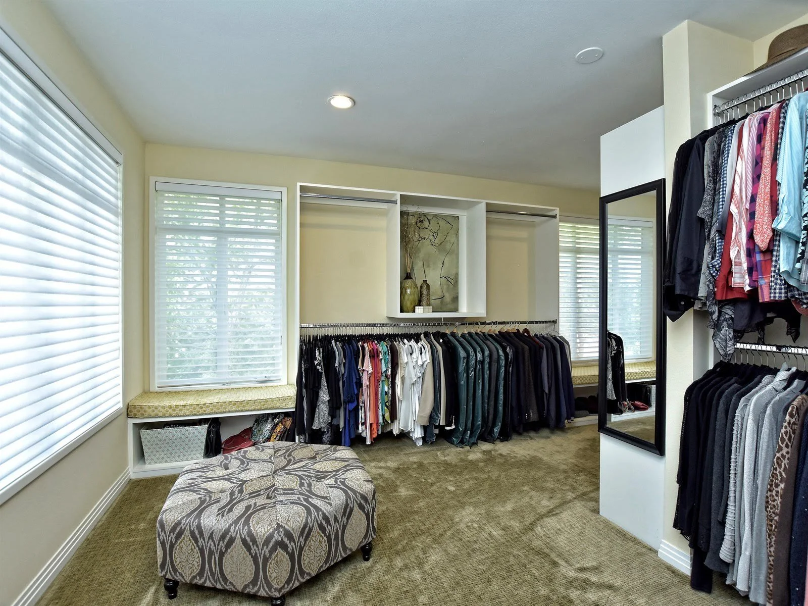 Spacious walk-in closet with clothes hanging on racks, a patterned ottoman, and large windows with blinds.