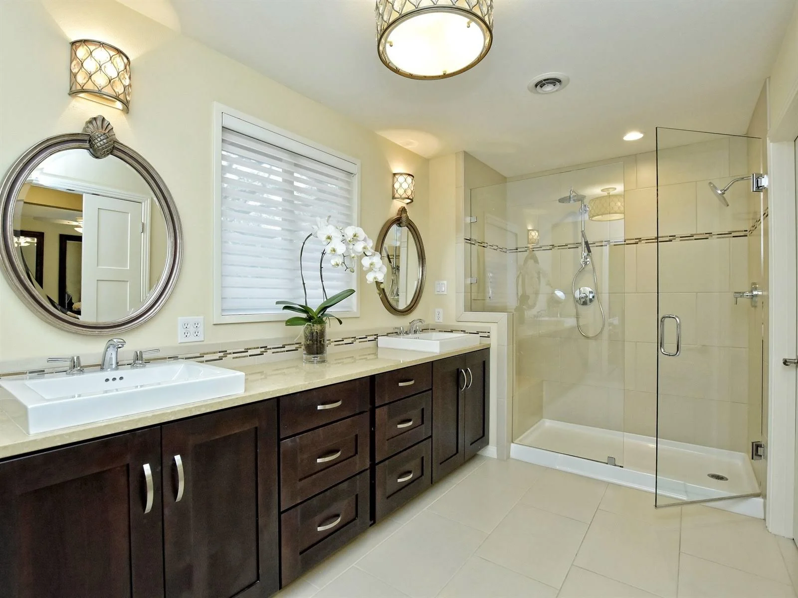Modern bathroom with double sinks, dark wood cabinetry, round mirrors, glass-enclosed shower, and orchid decoration.
