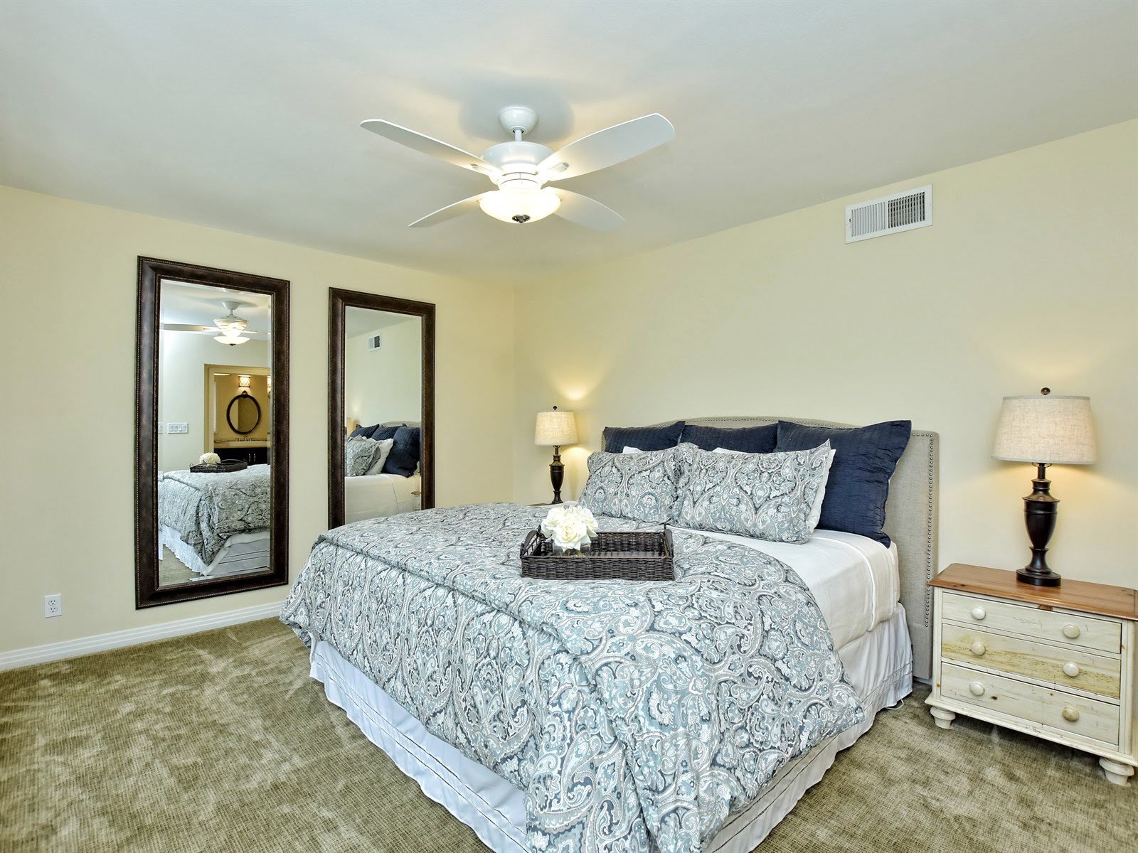 A bedroom with a bed, decorative bedding, two large mirrors on the wall, a ceiling fan, and a bedside table with a lamp.