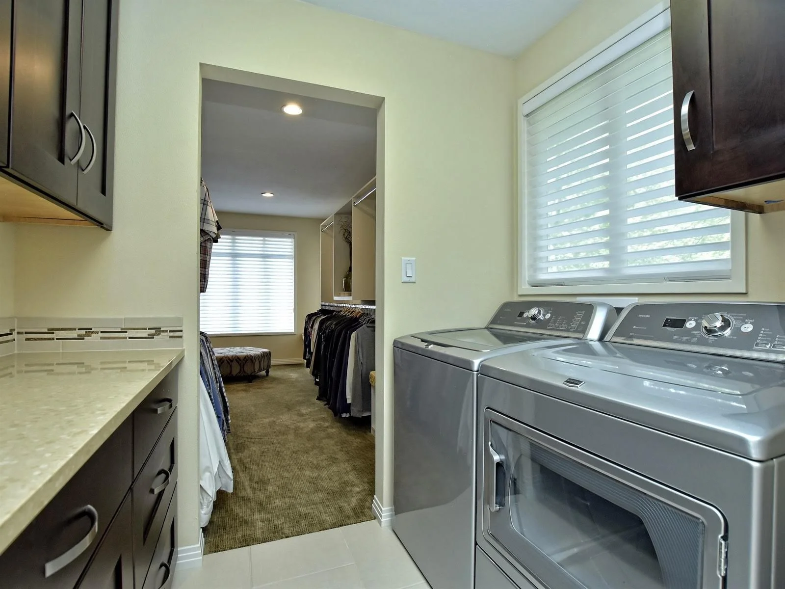 Laundry room with stainless steel washer and dryer, dark wood cabinets, and a view into an adjacent walk-in closet filled with hanging clothes.