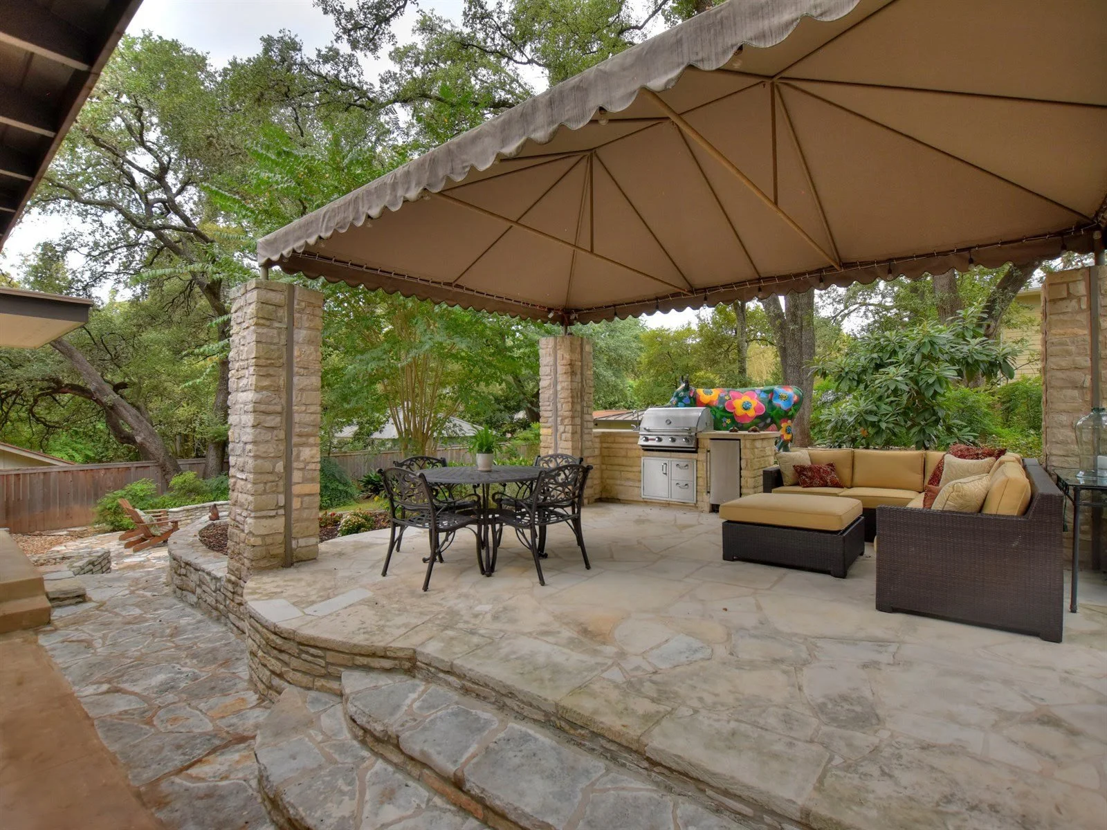 Outdoor patio with covered canopy, stone columns, dining table, sofa, barbecue grill, and colorful cow sculpture, surrounded by trees and garden.