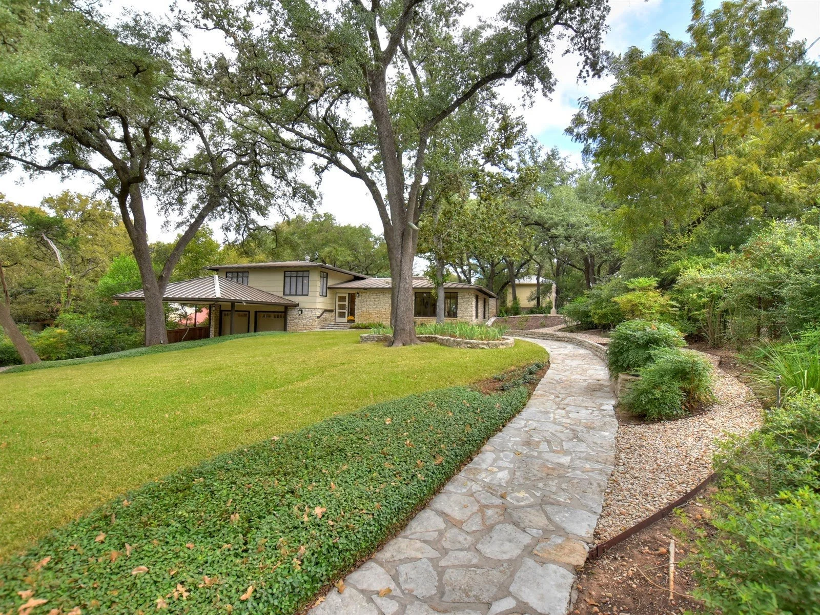 A modern house with stone exterior surrounded by trees and greenery, featuring a large, well-manicured lawn and a winding stone pathway leading to the entrance.