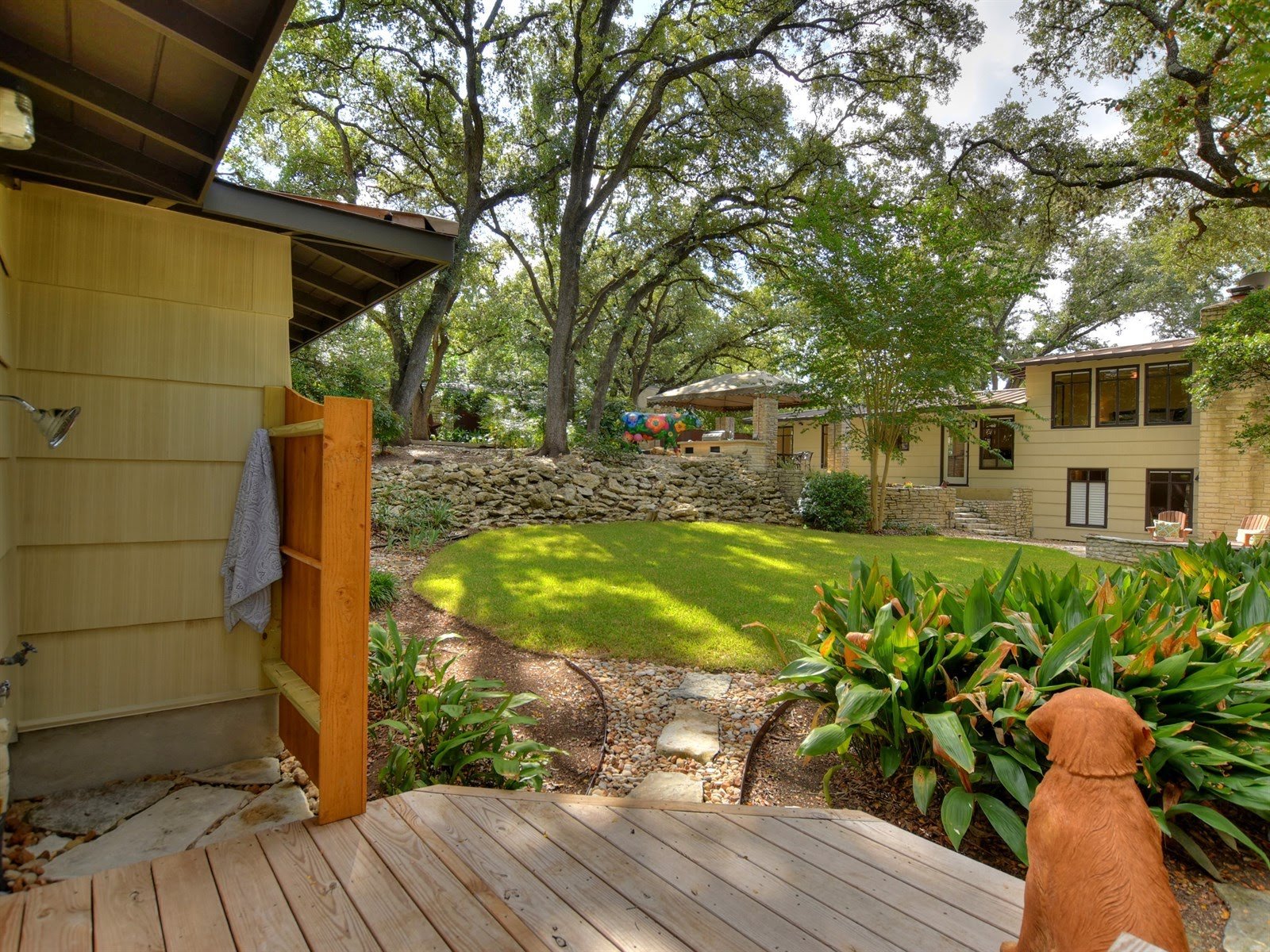 Backyard view with shower and dog statue, surrounded by lush greenery, a yellow house, and a stone pathway.
