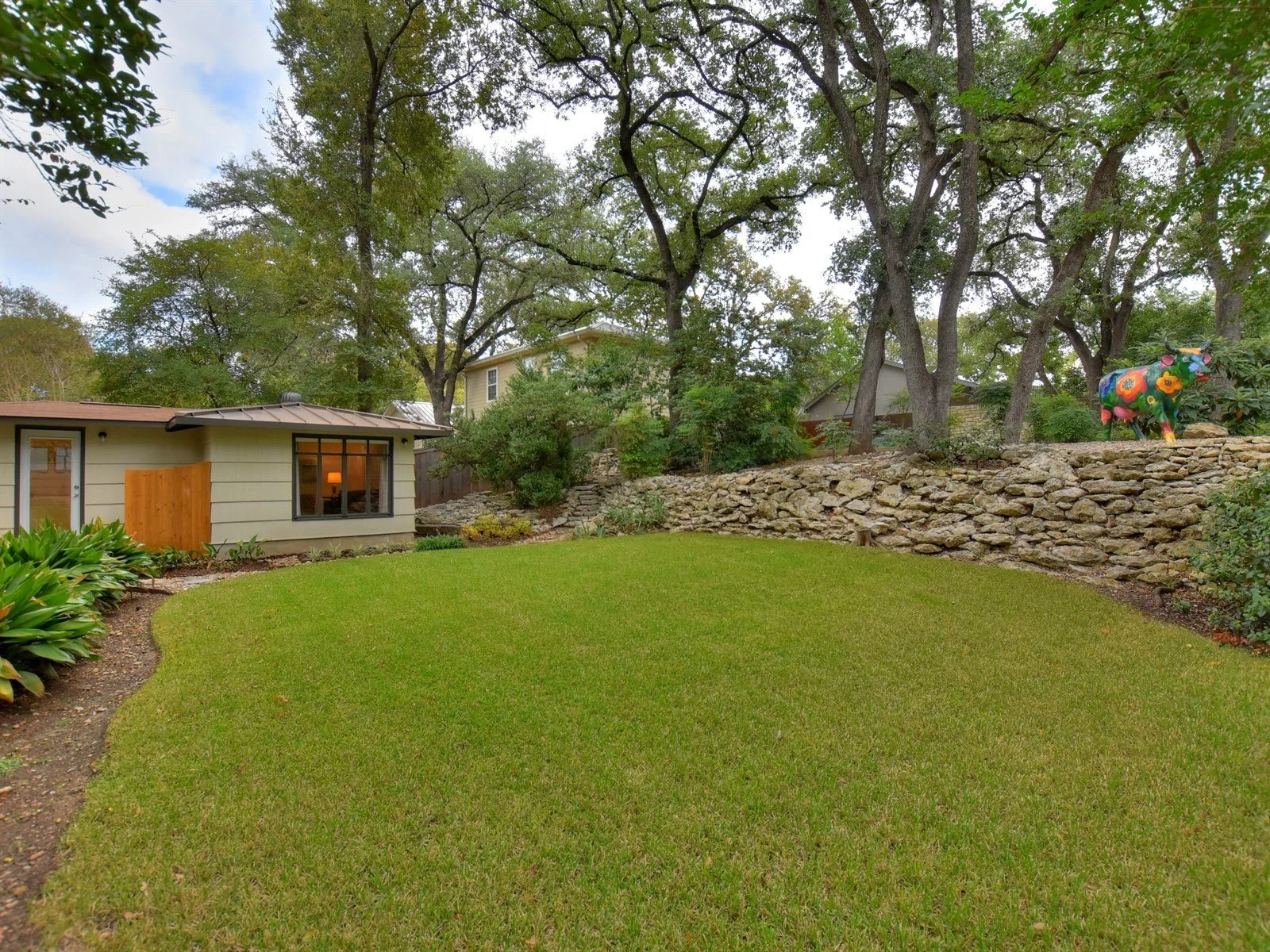 A lush green backyard with trimmed grass, trees, a stone retaining wall, and a house in the background. A colorful cow statue is on top of the wall.