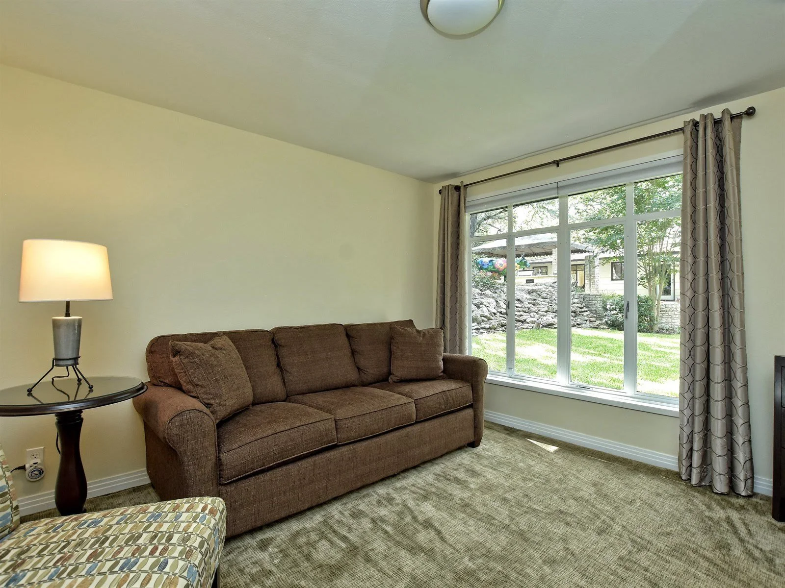 Living room with a brown sofa, side table with lamp, patterned armchair, large window with curtains, and a view of a green garden.