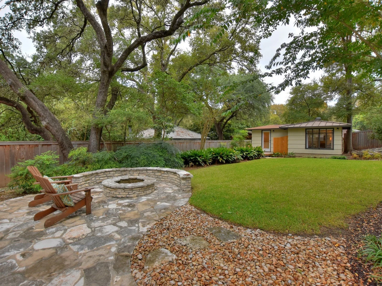 Backyard with stone patio, fire pit, and Adirondack chairs surrounded by trees and lush greenery. A small house or shed is visible in the background.