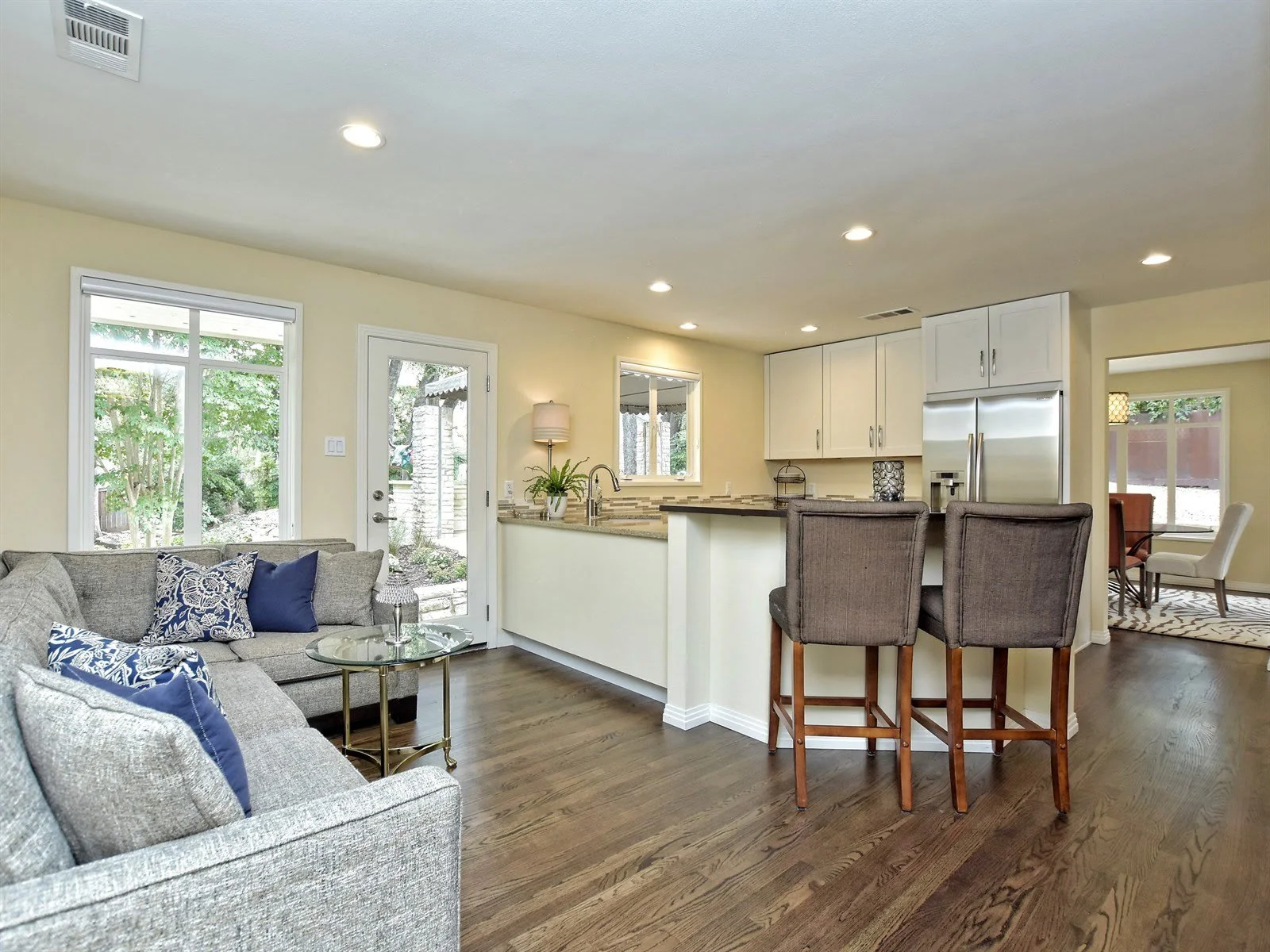 Open-concept living room and kitchen with hardwood floors, gray sectional sofa, patterned cushions, glass coffee table, countertop with barstools, stainless steel refrigerator, and dining area in the background.