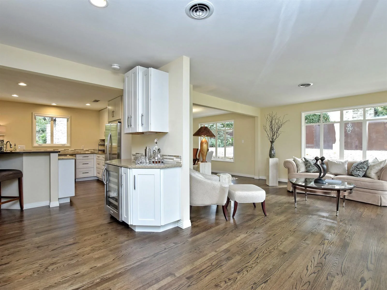 Open-concept living room and kitchen area with wooden flooring, featuring a sofa and armchairs, a coffee table, kitchen counters, and large windows providing natural light.
