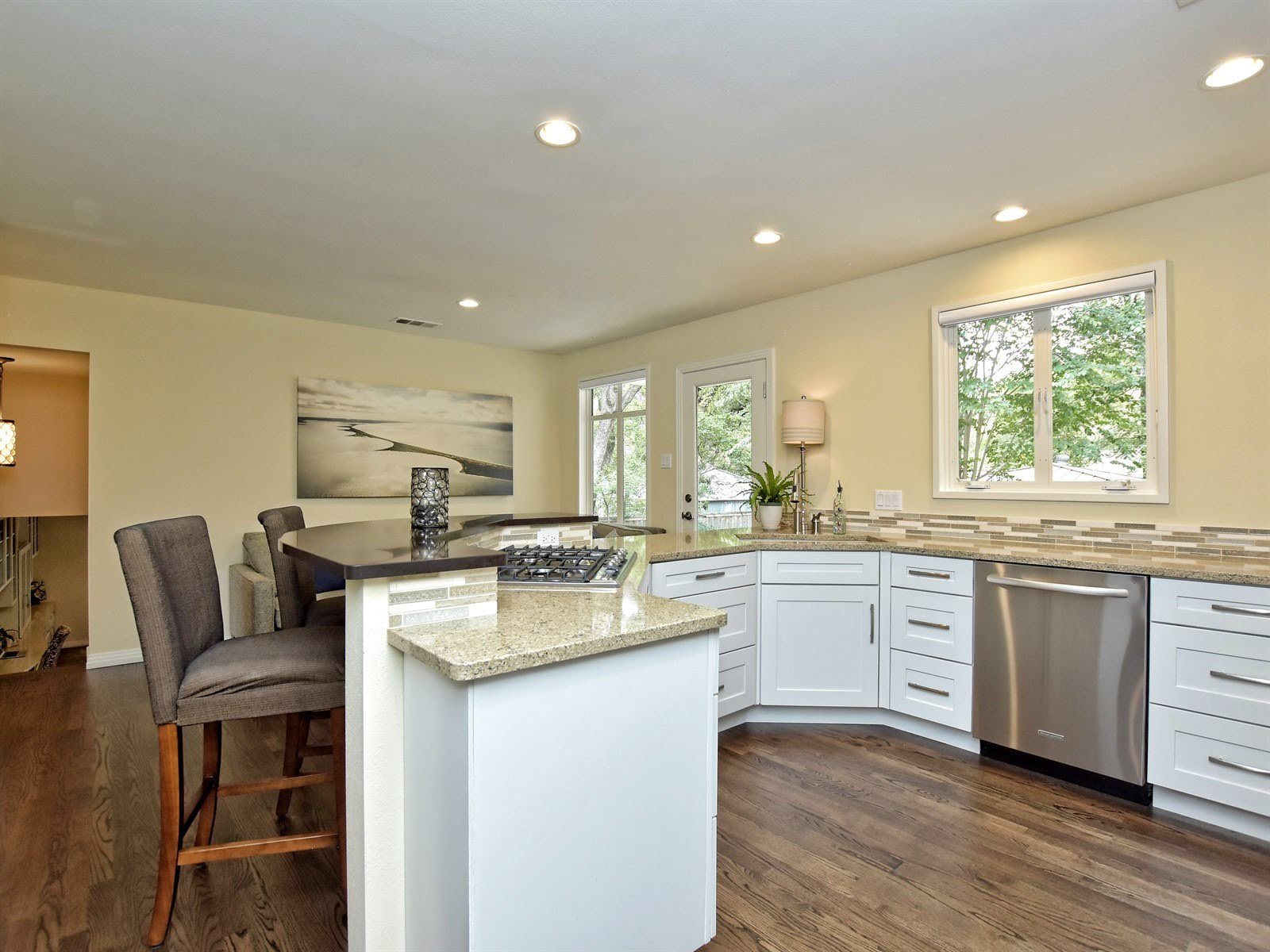 Modern kitchen with light wood flooring, white cabinets, stainless steel dishwasher, granite countertops, and a breakfast bar with high chairs. Large window and an open door provide natural lighting.