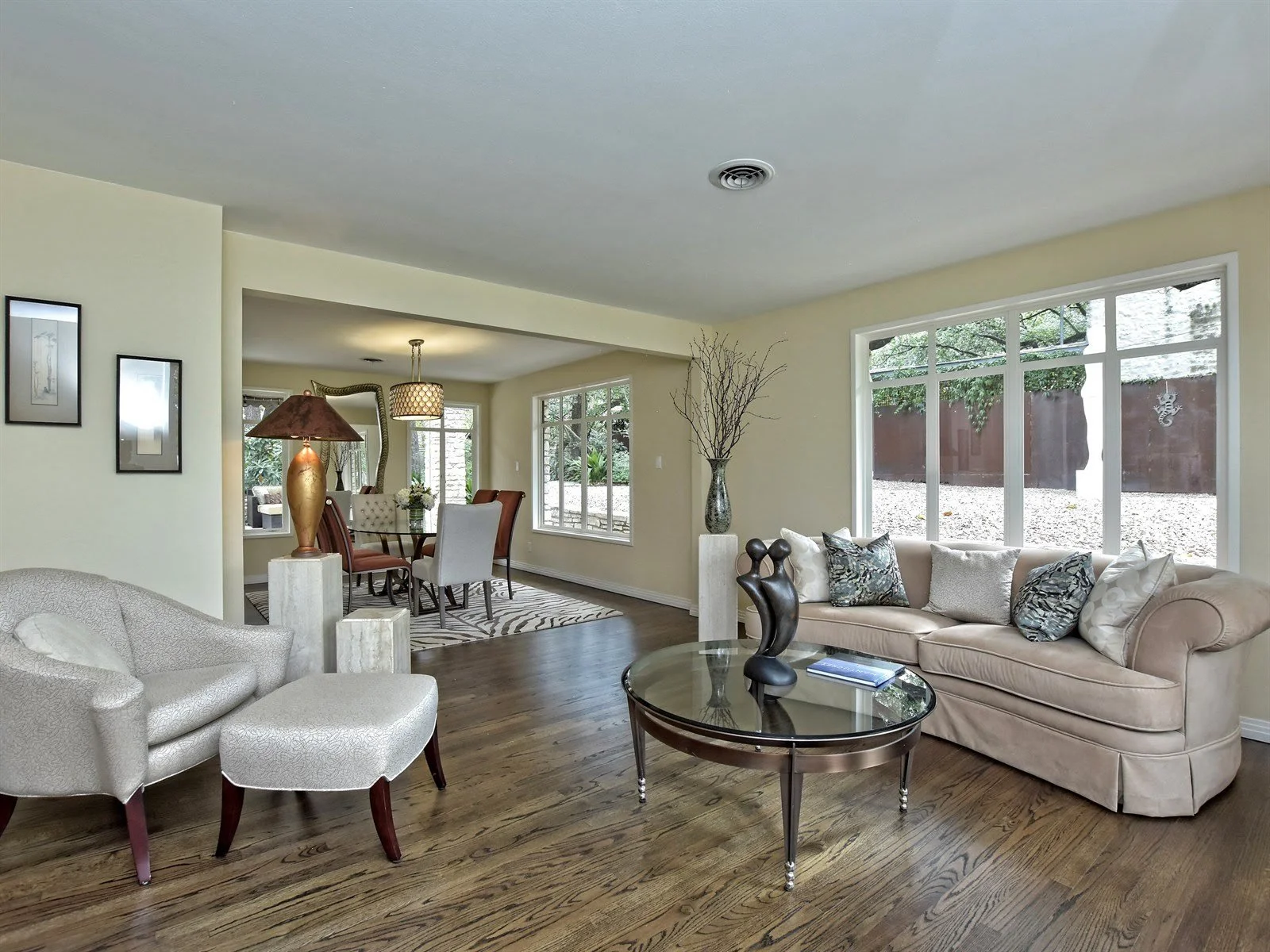 Spacious living room with beige walls, featuring a plush beige sofa with decorative pillows, glass coffee table with a sculpture, and an armchair with an ottoman. Large windows provide natural light and a view of the outside. Adjacent dining area wit