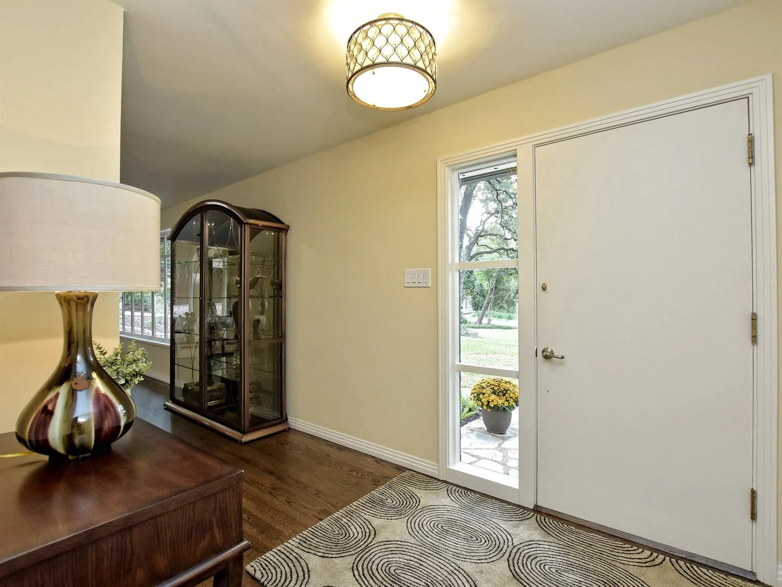 Entryway with a white door, glass windows, wooden flooring, and a cabinet displaying items. A modern lamp is on a dark wooden table, and there's a patterned rug on the floor. Outside view includes greenery and flowers.