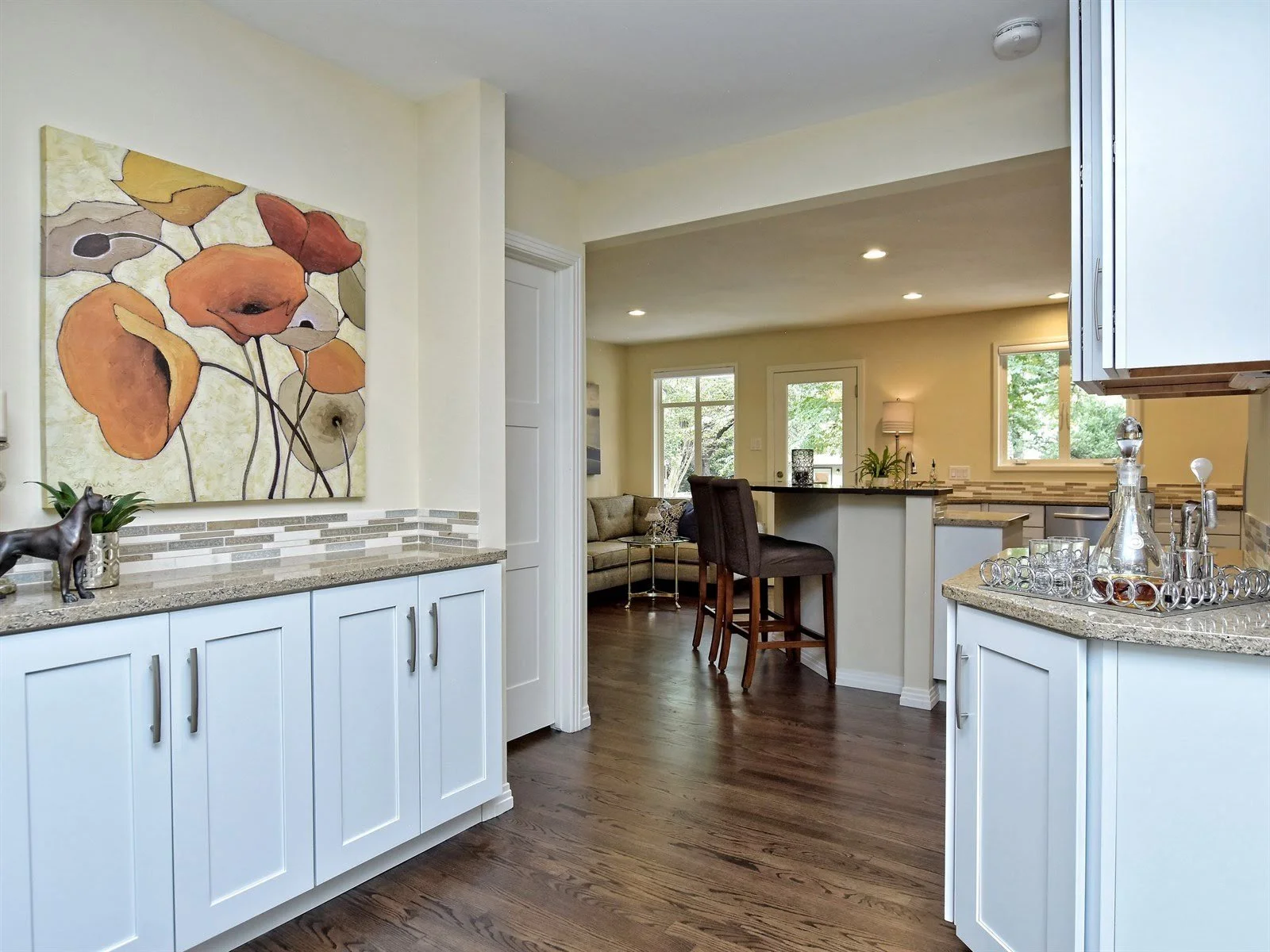 Modern kitchen and dining area featuring white cabinets, dark wooden flooring, and a colorful abstract painting on the wall. The kitchen counter has glassware and a decorative sculpture. Bar stools are positioned at the kitchen island, and a living r