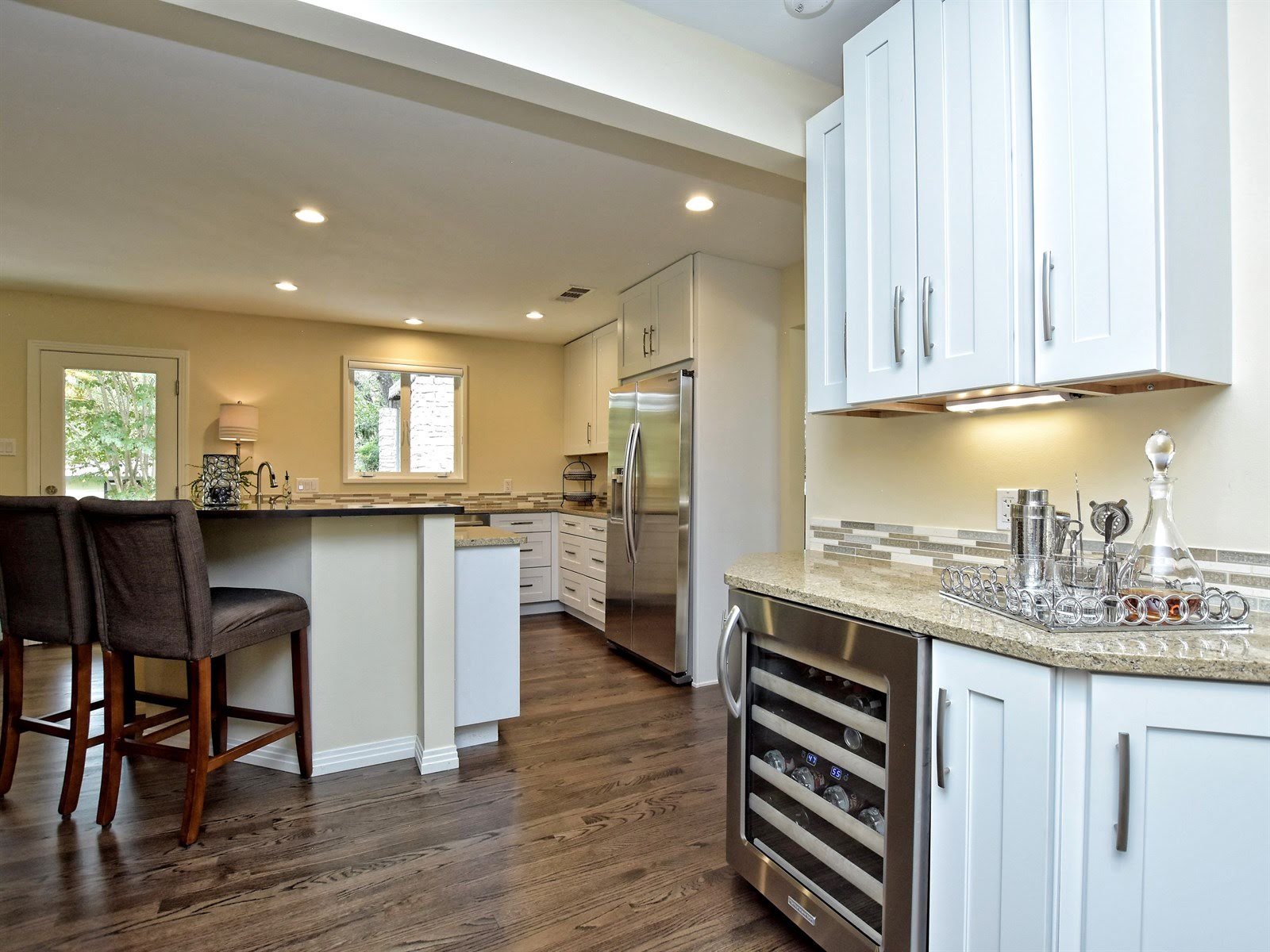 Modern kitchen with hardwood floors, stainless steel appliances, white cabinets, and a wine cooler. A countertop features a tray with bar accessories.