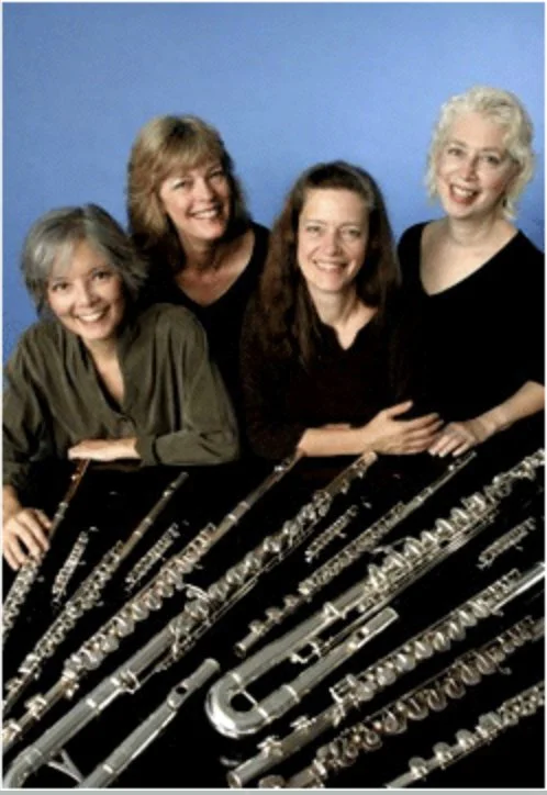 Four women smiling behind a table of silver flutes and musical instruments against a blue background.