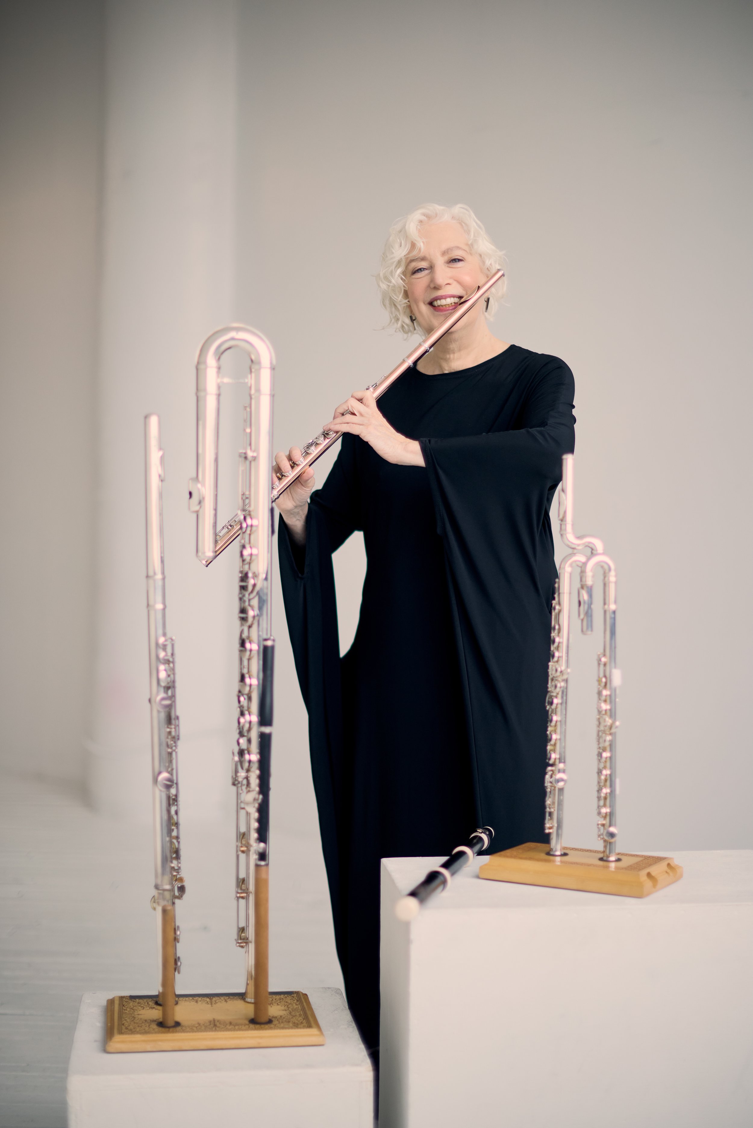 Wendy Stern, performer and educator, dressed in black dress, holding 14k rose gold Brannen Bros. flute. There is an alto flute, bass flute, piccolo, Baroque flute, a curved headjoint flute, and a Jupiter Waveline flute in the foreground